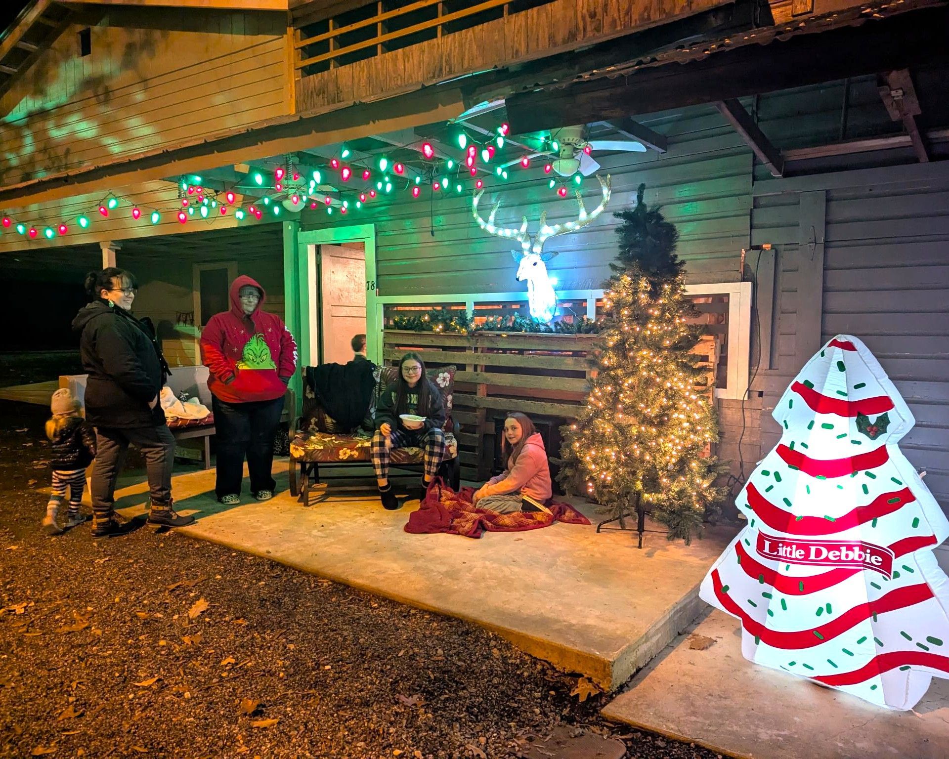 People sitting outside a decorated home at night beside a lit Christmas tree and glowing snowman decoration
