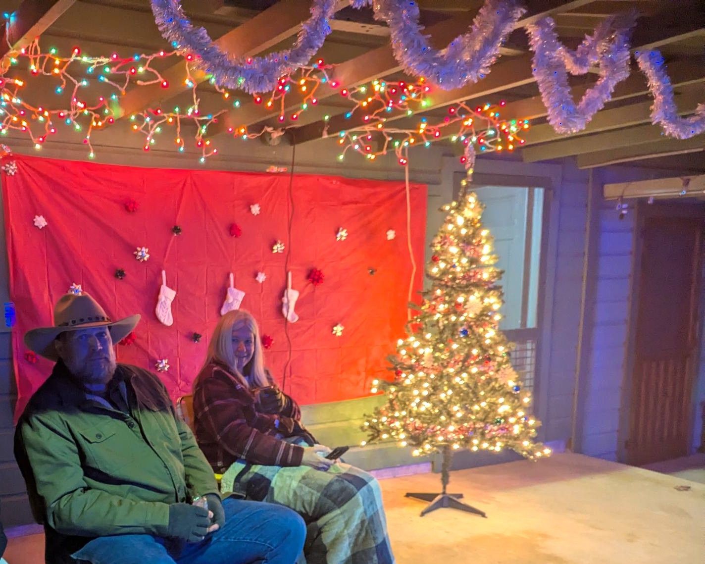 Two people seated under holiday lights beside a decorated Christmas tree and red backdrop.