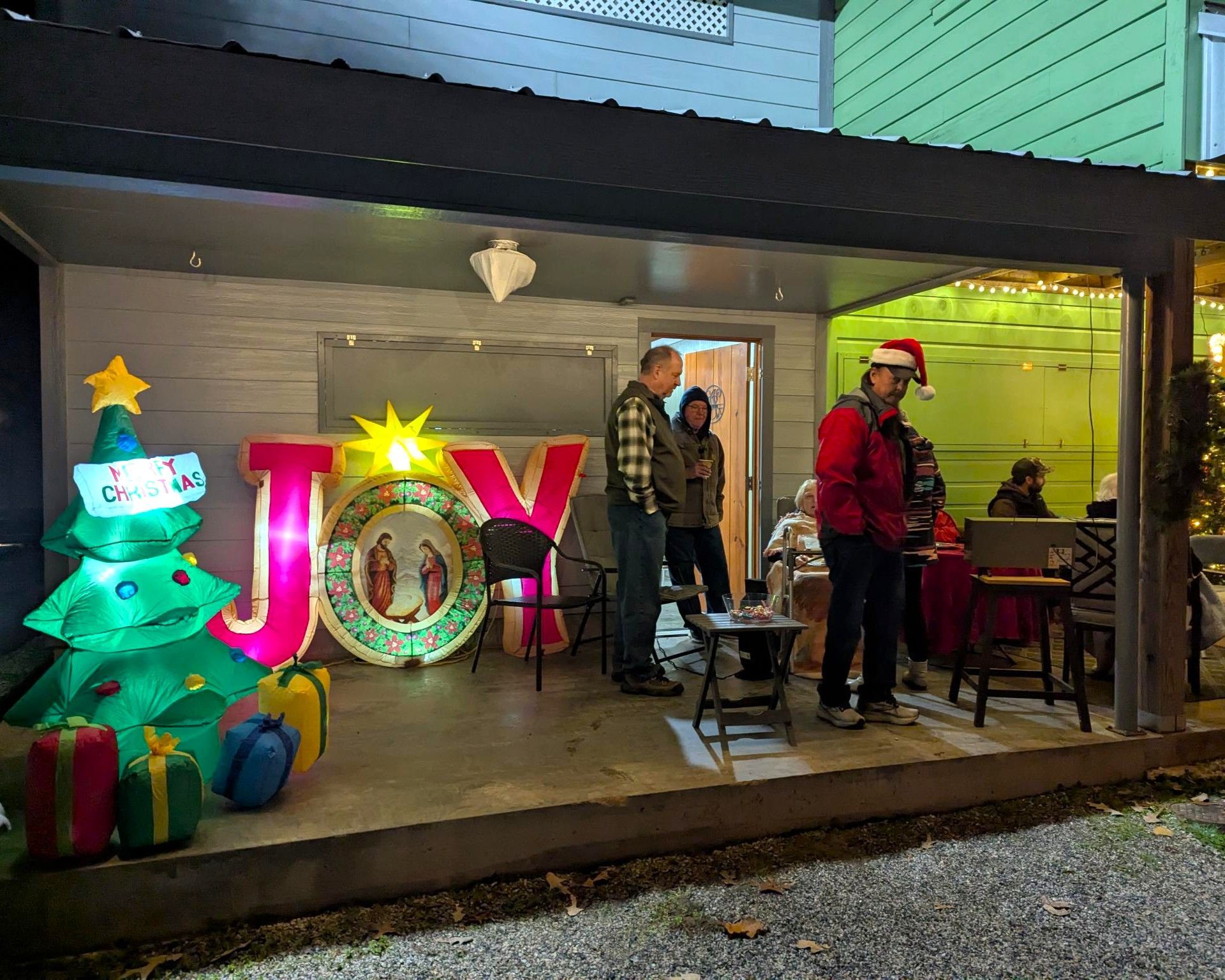 Holiday porch display with glowing Christmas tree, large “JOY” sign, gifts, and two people standing by the house