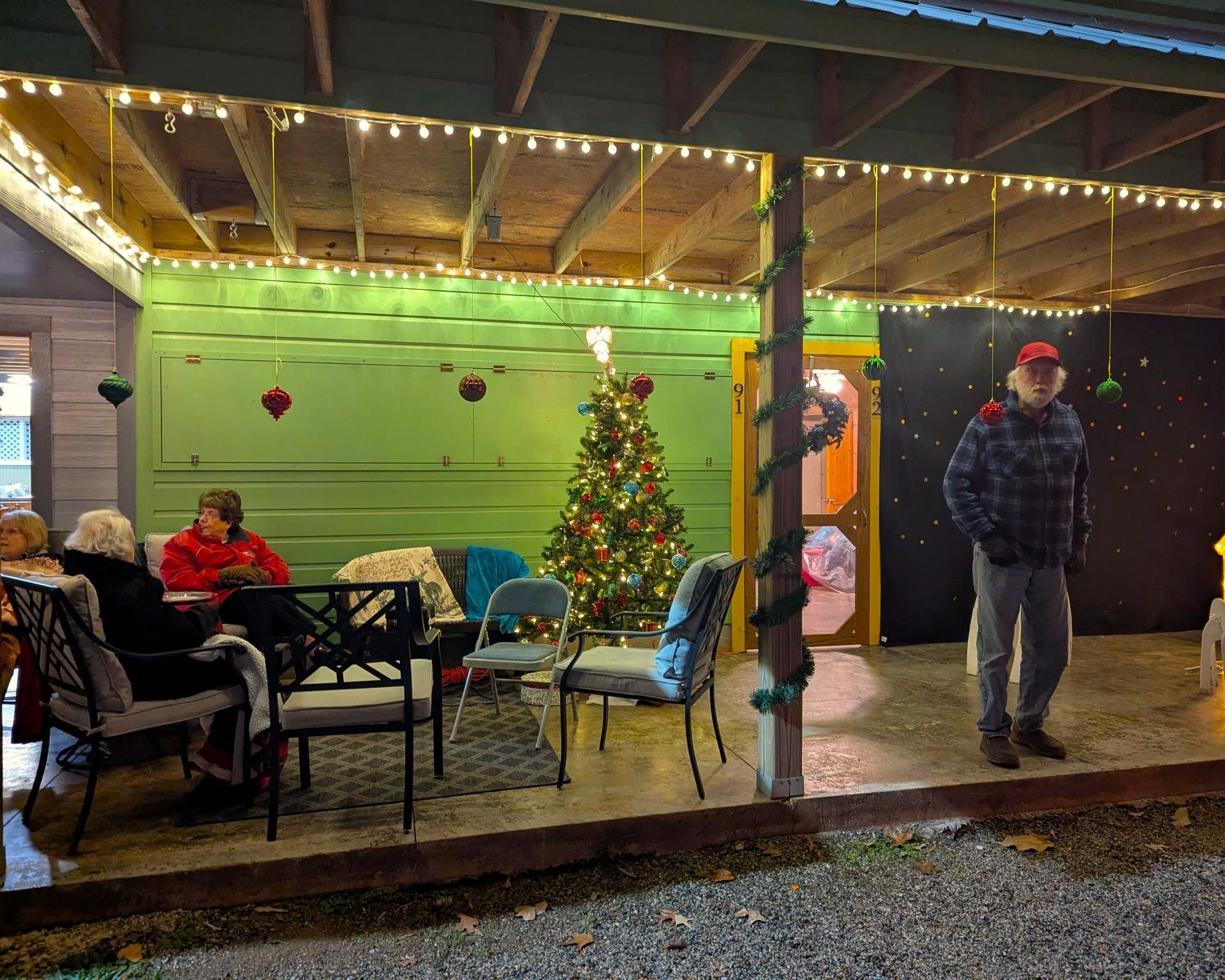 Covered porch with string lights, Christmas tree, chairs, and two people standing outside at night.