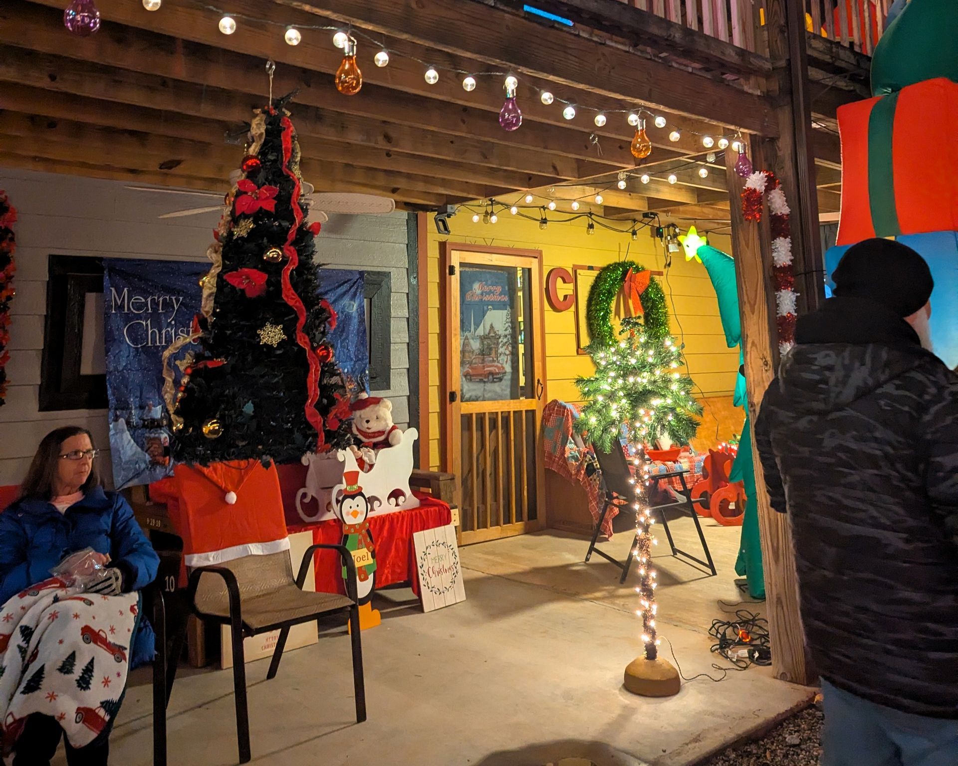Festive porch with Christmas trees, red decorations, and people gathered under string lights