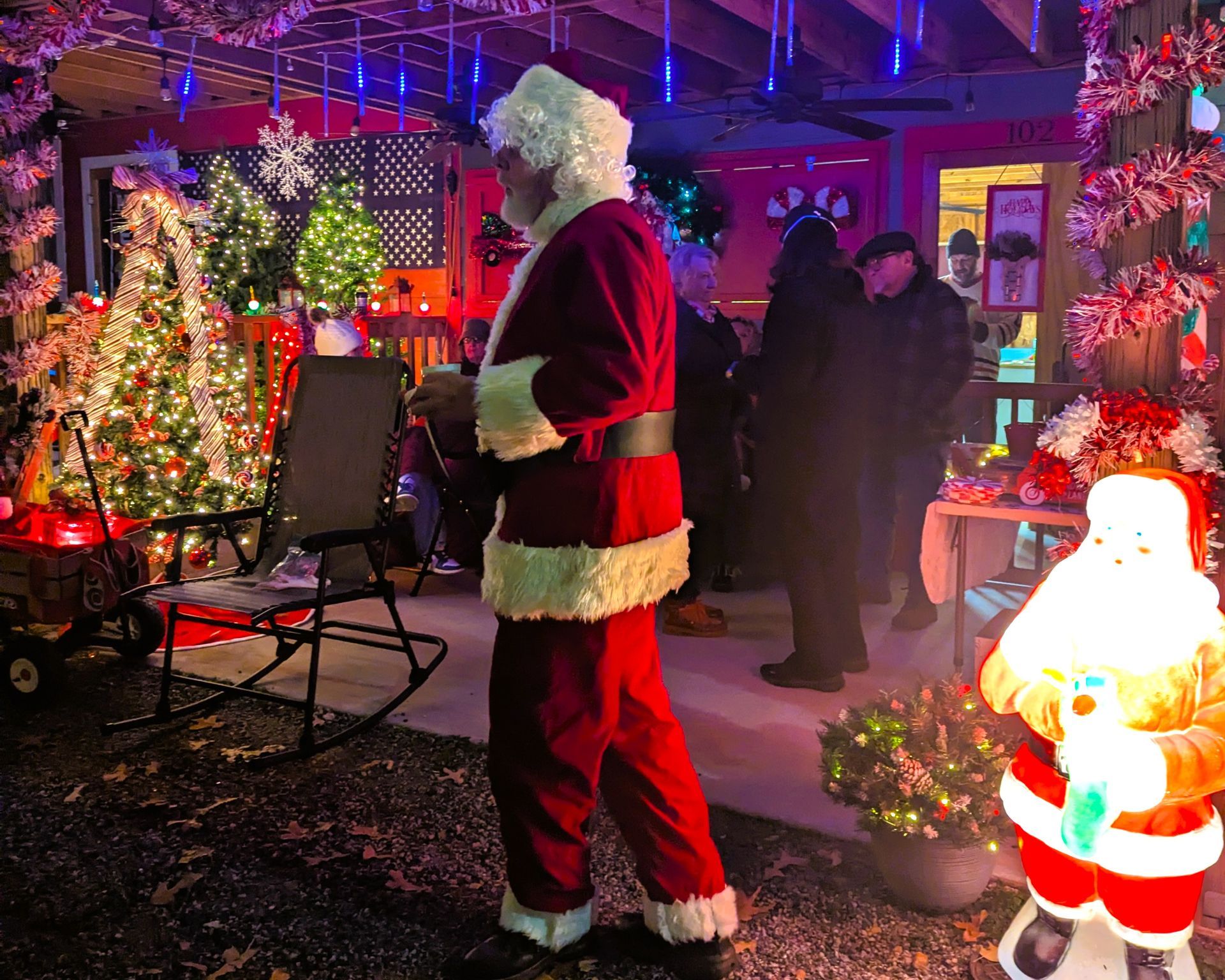 Santa in a red suit stands outside a festive Christmas display with colorful lights and decorations.
