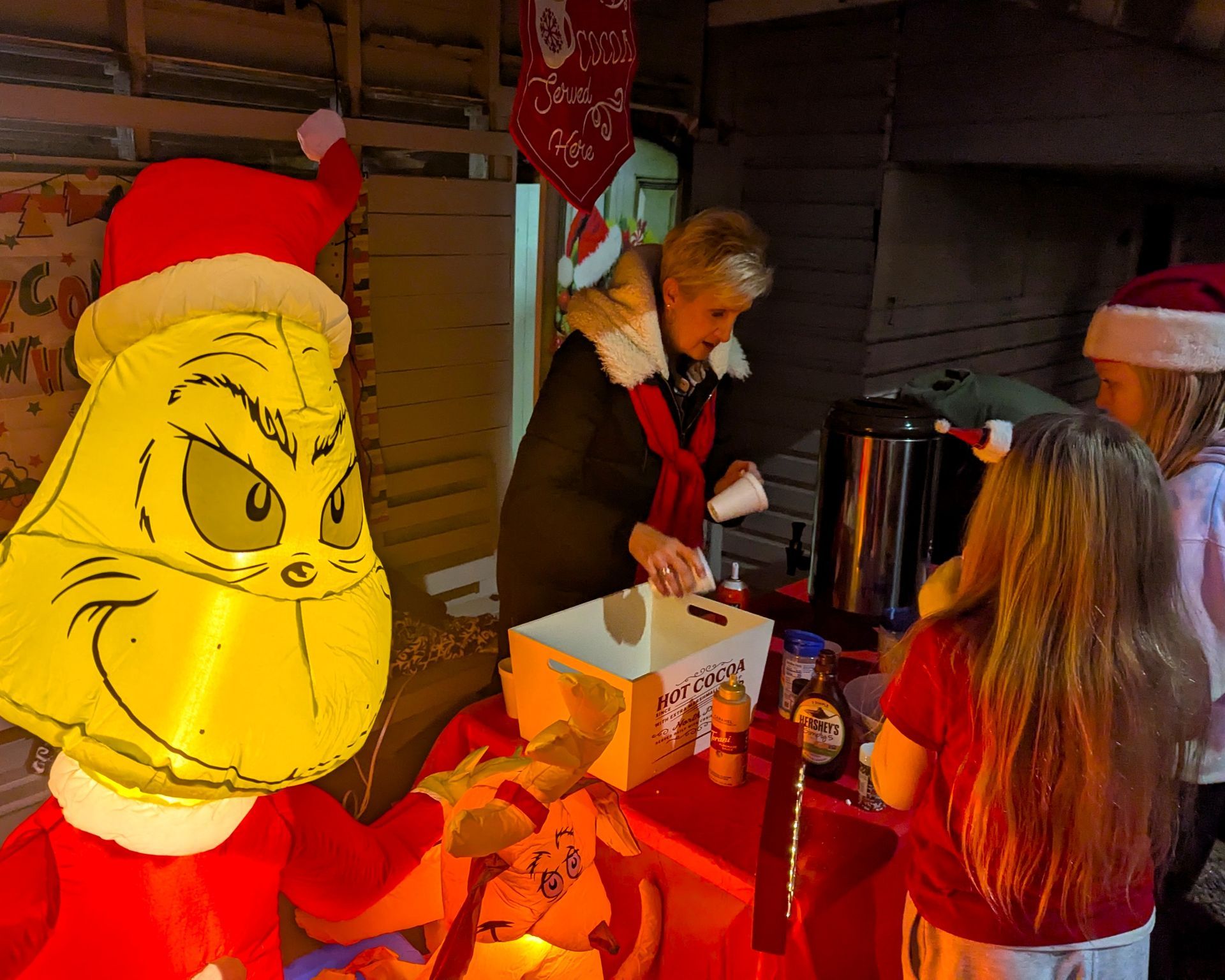 Grinch character at a festive table serving drinks beside two people in a warm holiday setting