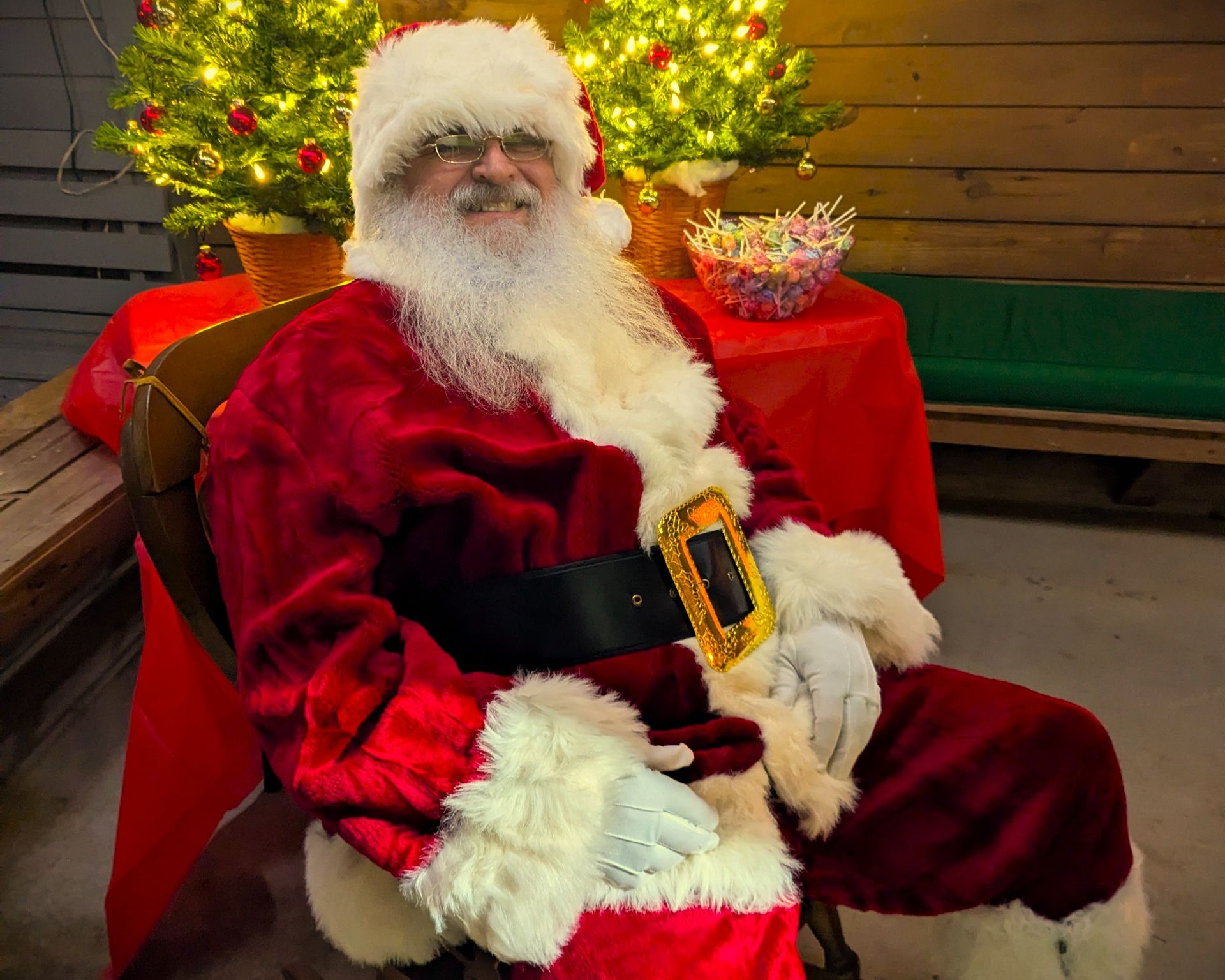Santa Claus in a red suit sitting by a lit Christmas tree, smiling and holding a gift basket.