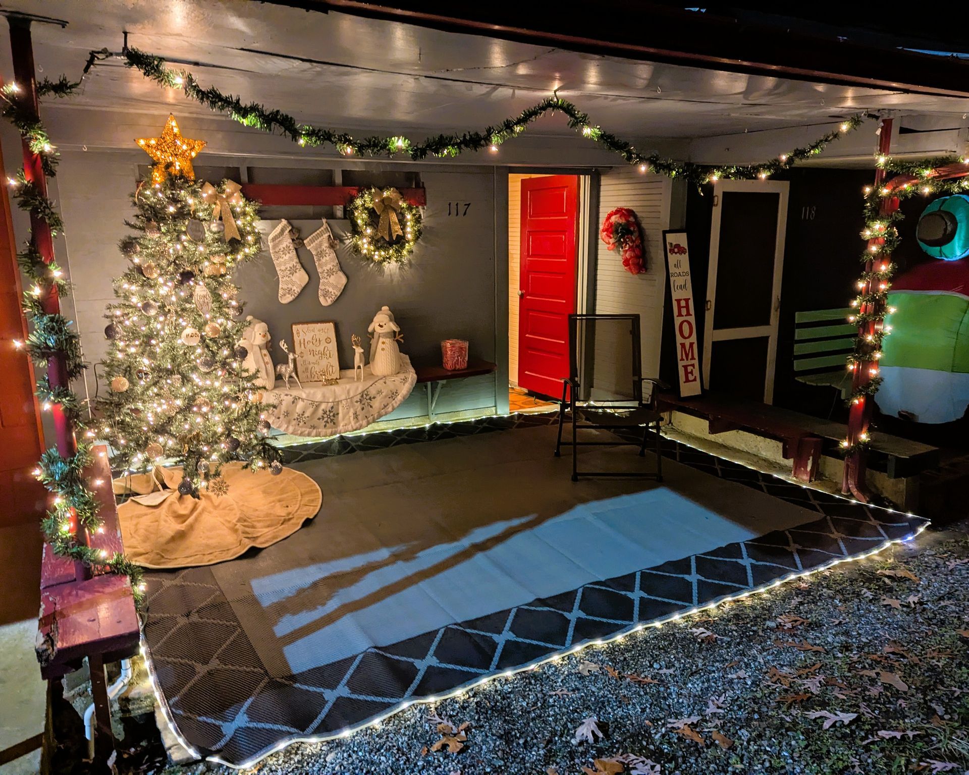 Holiday-decorated patio with Christmas tree, lights, stockings, and a glowing red door at night