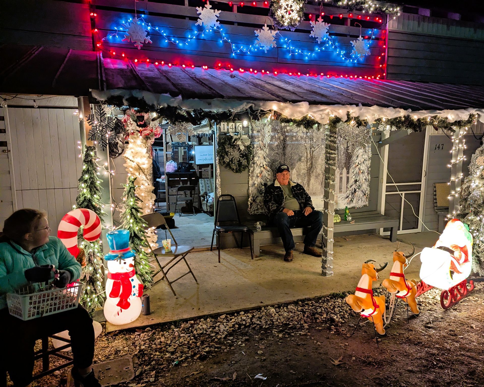 House decorated for Christmas with lights, garlands, snowmen, and people sitting on the porch at night