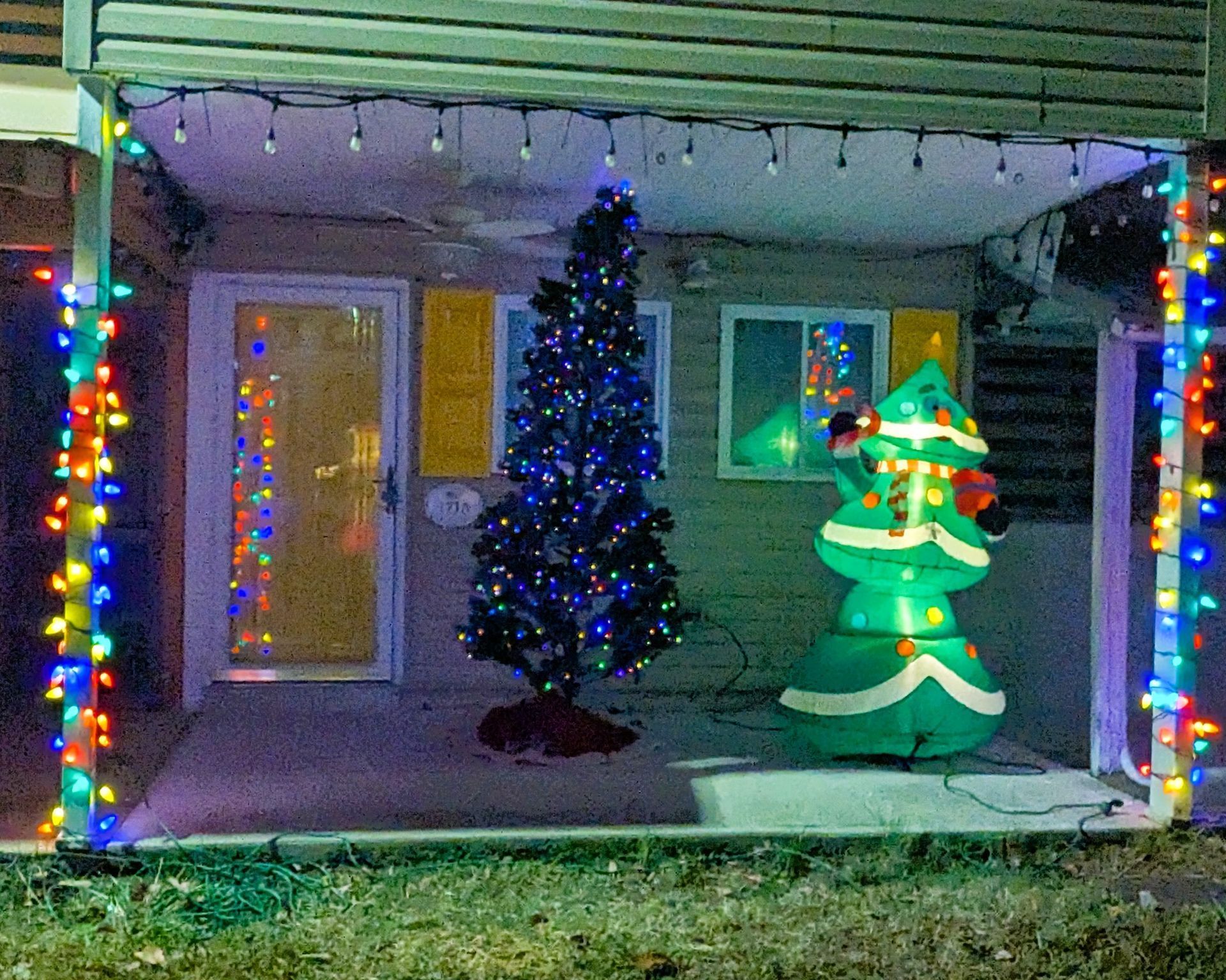House porch decorated for Christmas with colorful lights, a lit tree, and an inflatable holiday character.