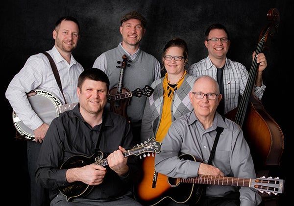 Six musicians posing with banjos, guitar, and bass against a dark background