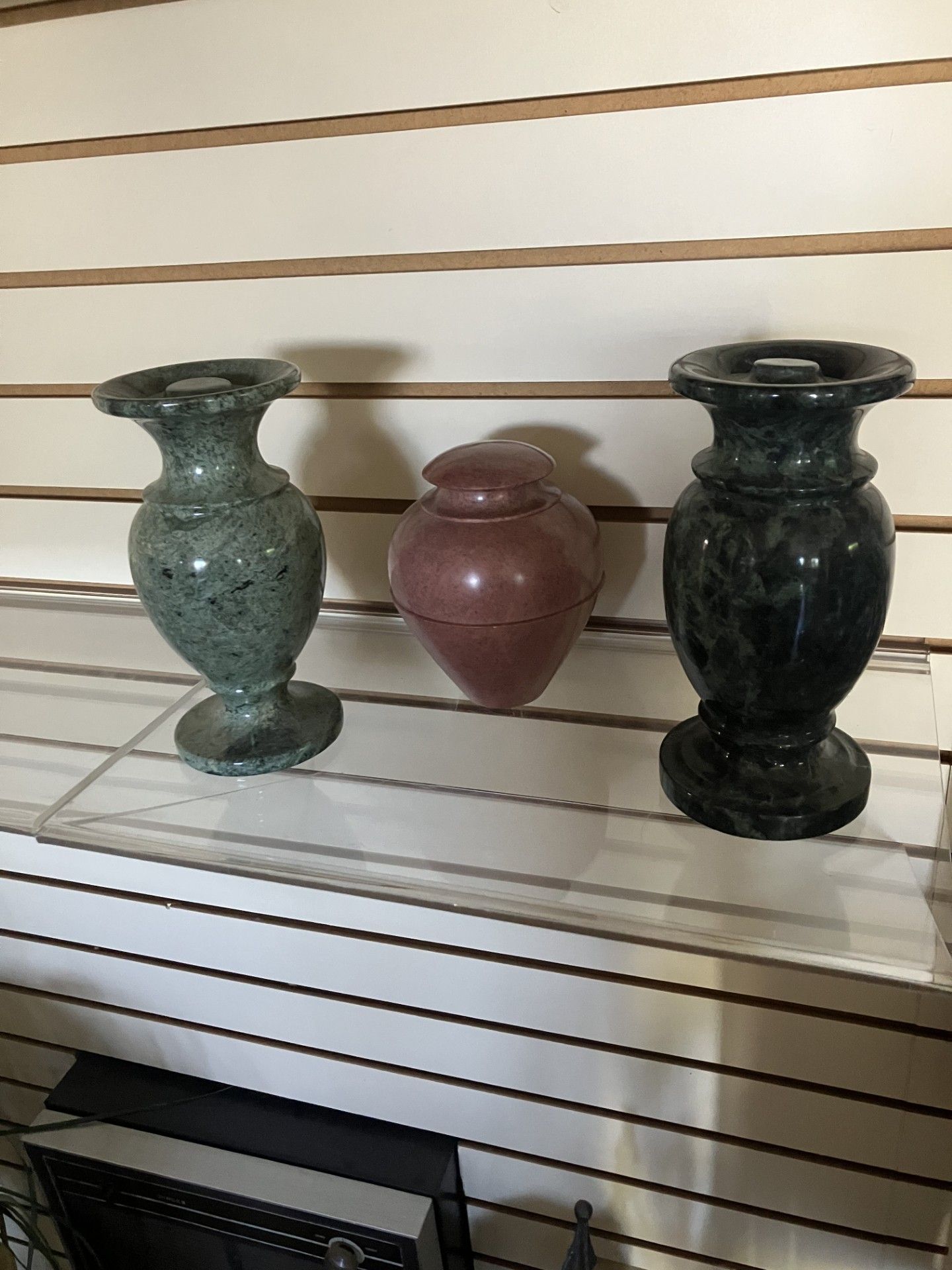 Three decorative vases on a slatted display shelf, in green, mauve, and dark gray. Three decorative vases on a slatted display shelf, in green, mauve, and dark gray.