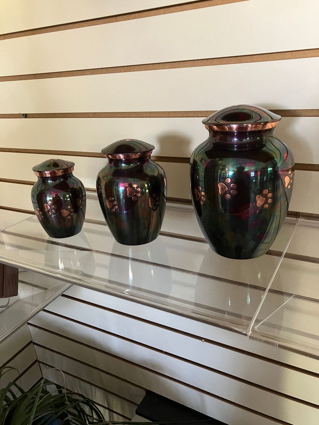 Three dark glass urns displayed on a white slatwall shelf, with the largest on the right.