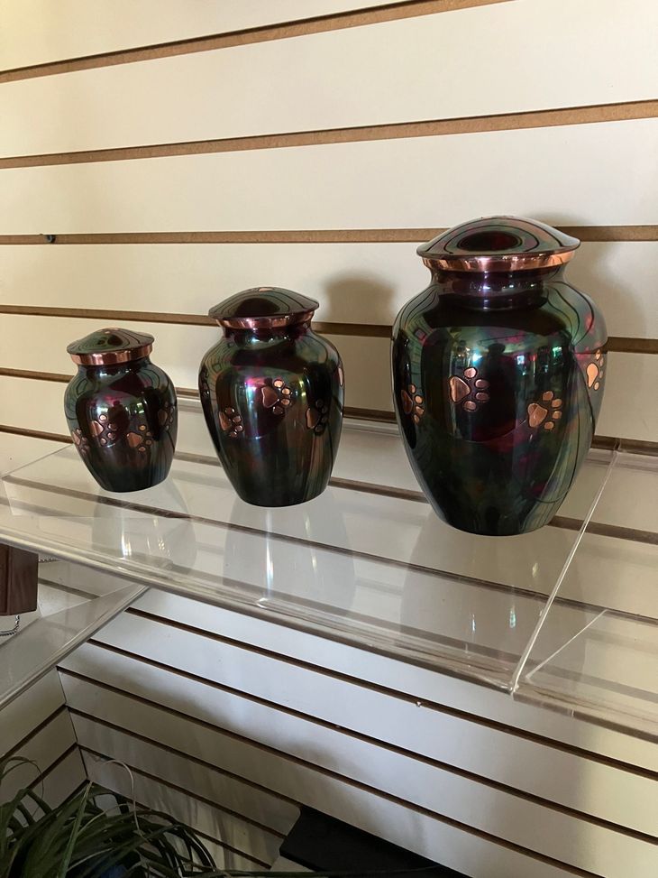 Three dark glass urns displayed on a white slatwall shelf, with the largest on the right.