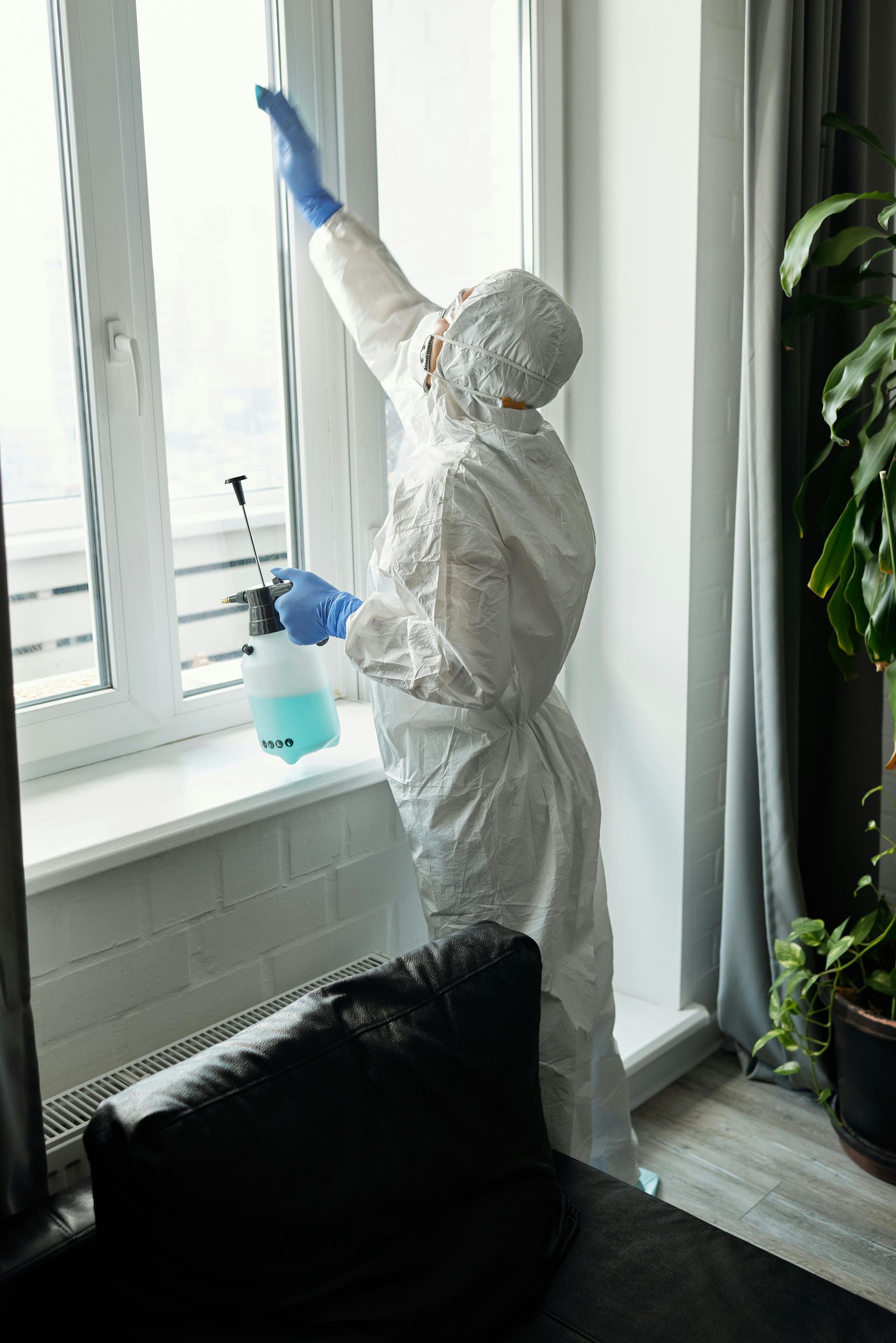 Person in hazmat suit spraying window, blue gloves, in room.