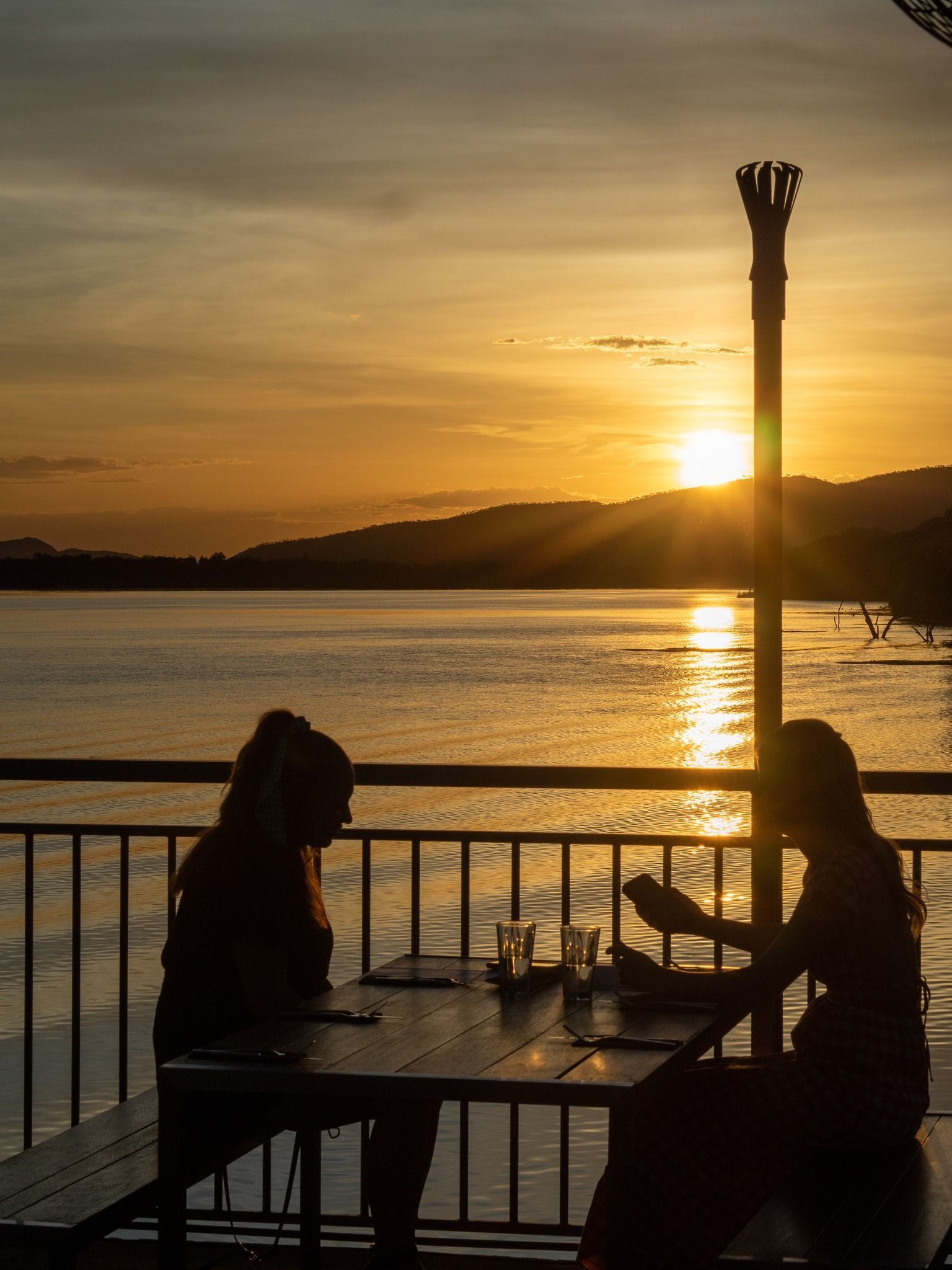 Two women are sitting at a table by the water at sunset.