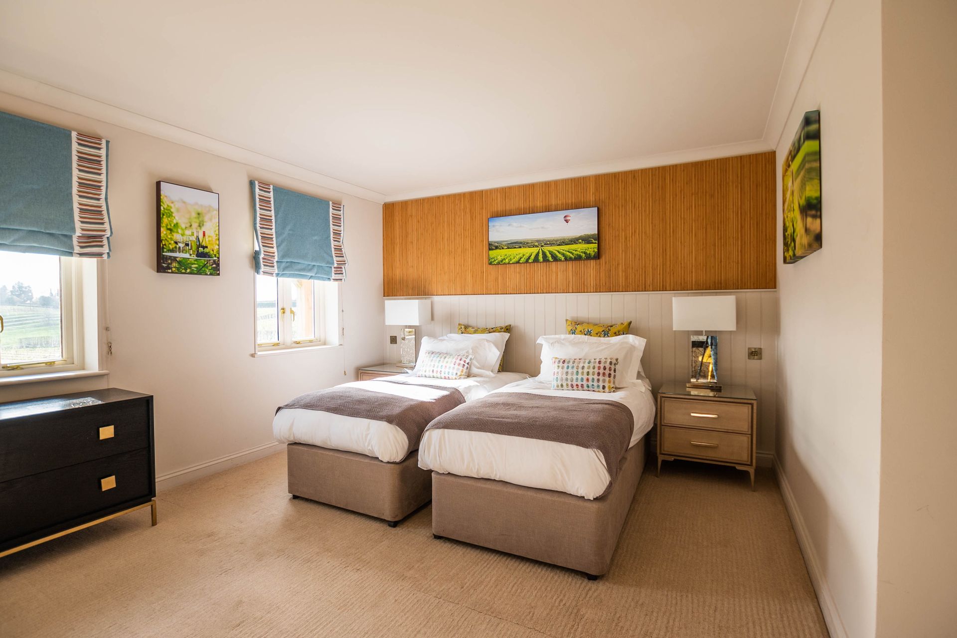 A hotel bedroom with two twin beds, a wooden headboard, and tan carpet. Artwork and lamps are on the bedside tables.