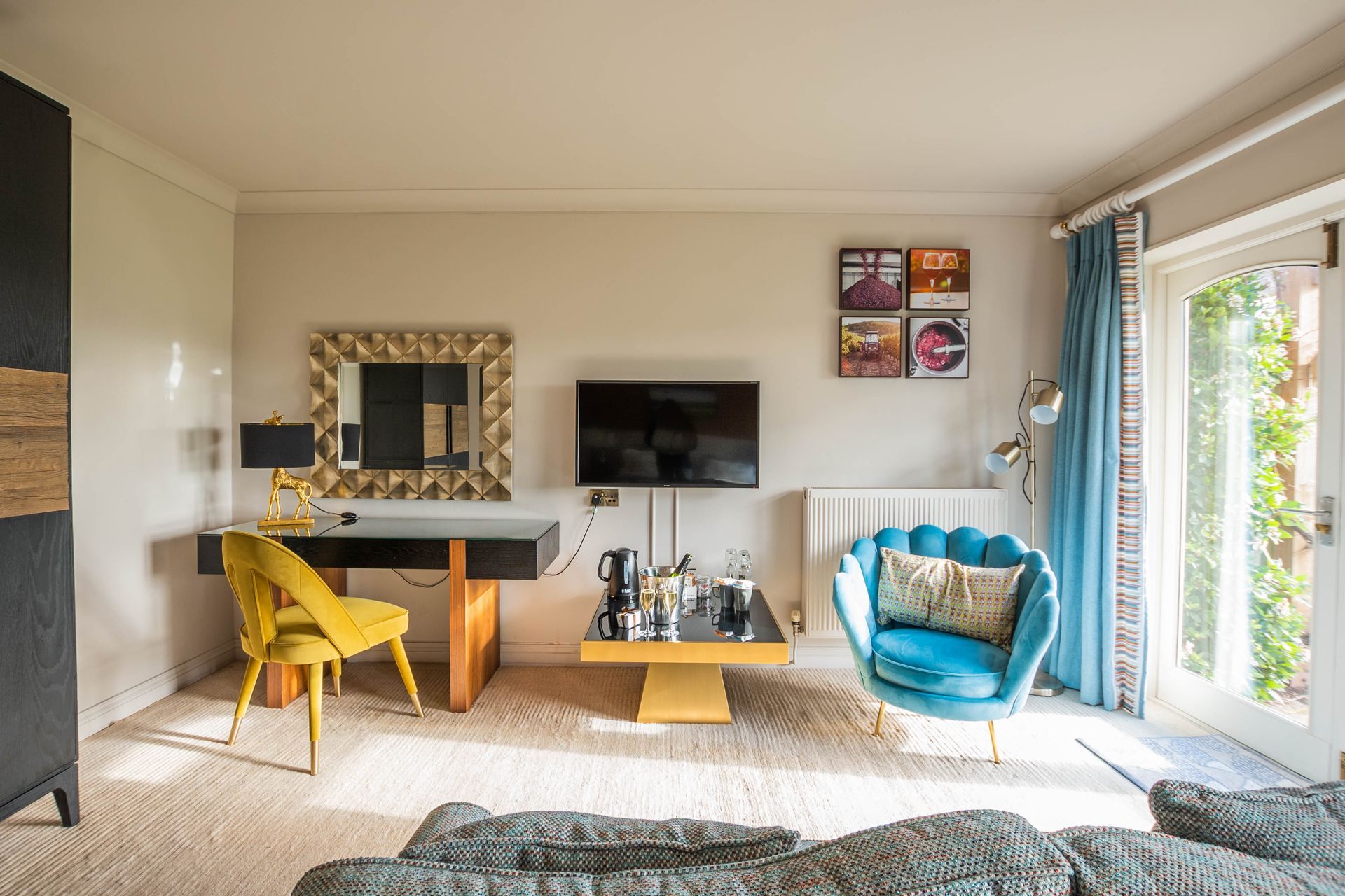 A living room with a desk, armchair, and TV. Yellow and blue accents pop against neutral walls and carpet, with a door to the right.