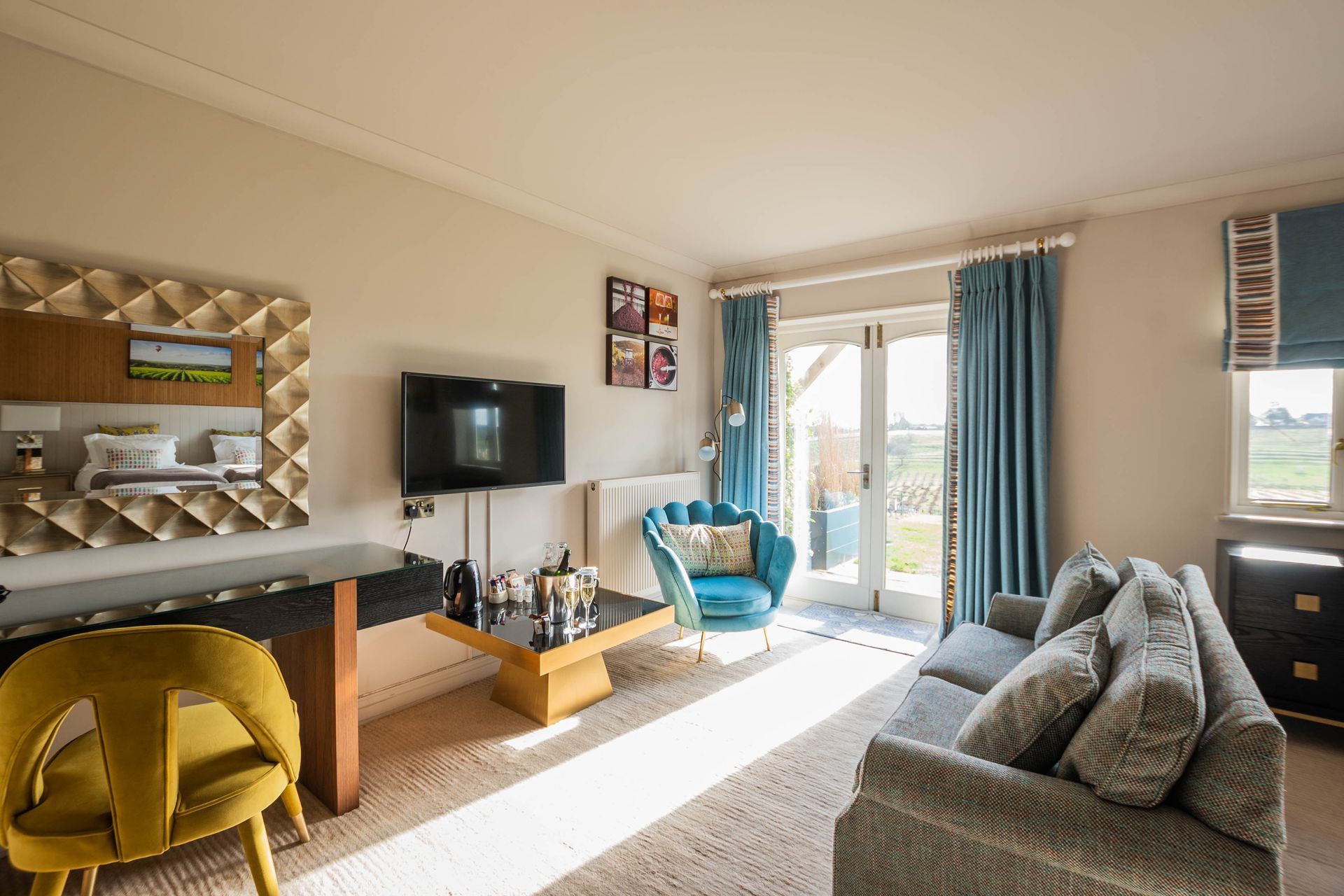 A brightly lit living room with a blue sofa, accent chair, and curtains. Features a desk with a gold chair, a TV, and a view of the outdoors.