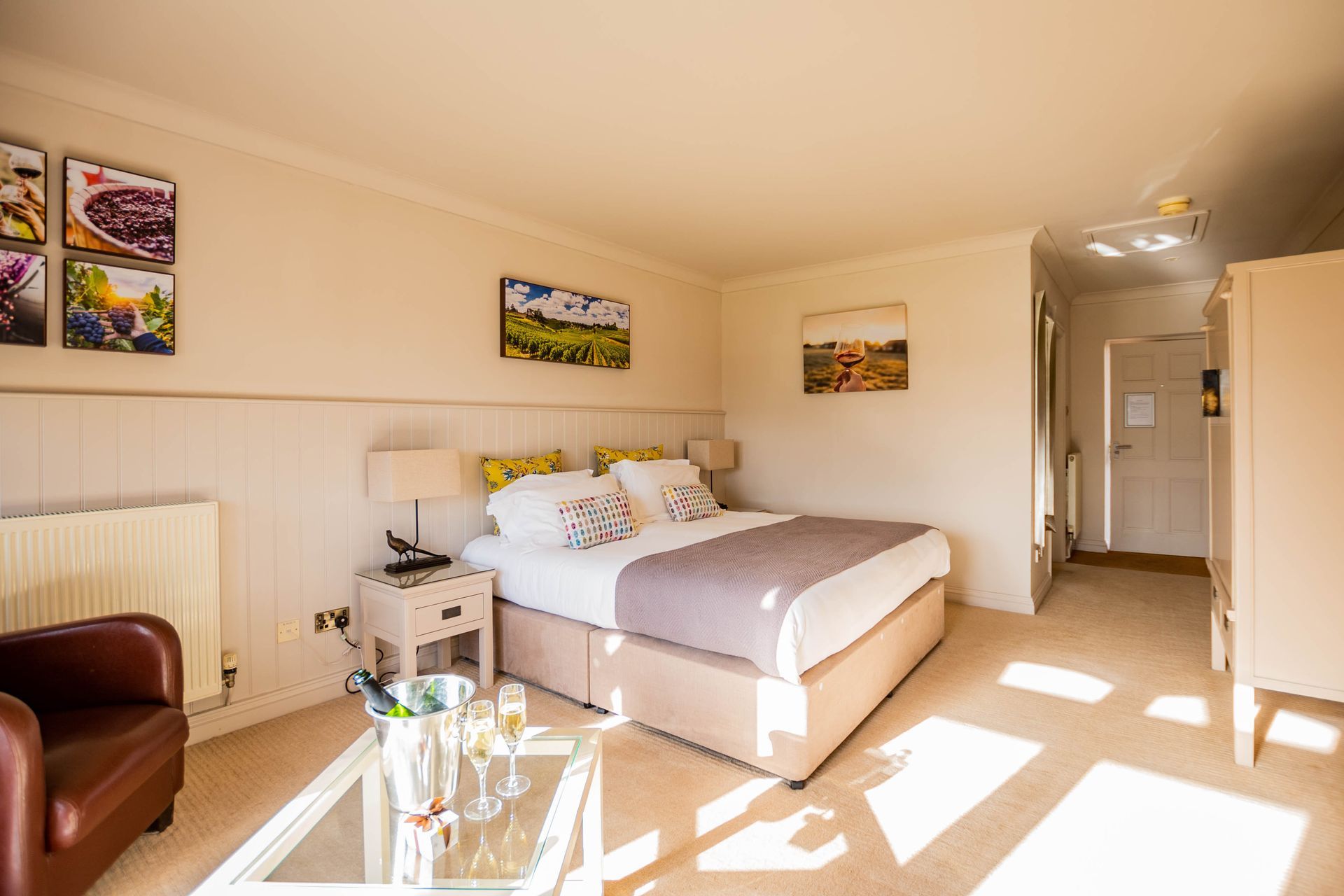 Bright bedroom with a double bed, arm chair, and bedside table. Champagne bucket on a glass table; sunlight streams in.
