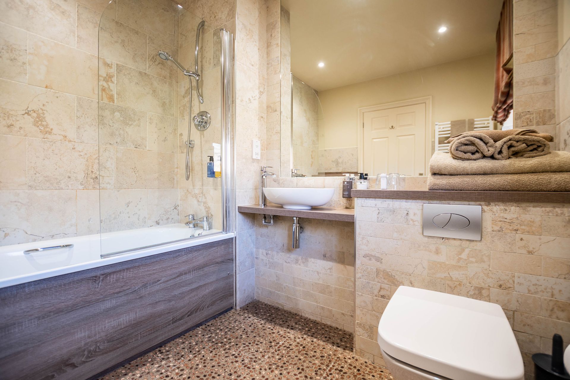 Bathroom with beige stone walls and a pebble floor. Features include a bathtub, sink, and toilet with a stack of towels.
