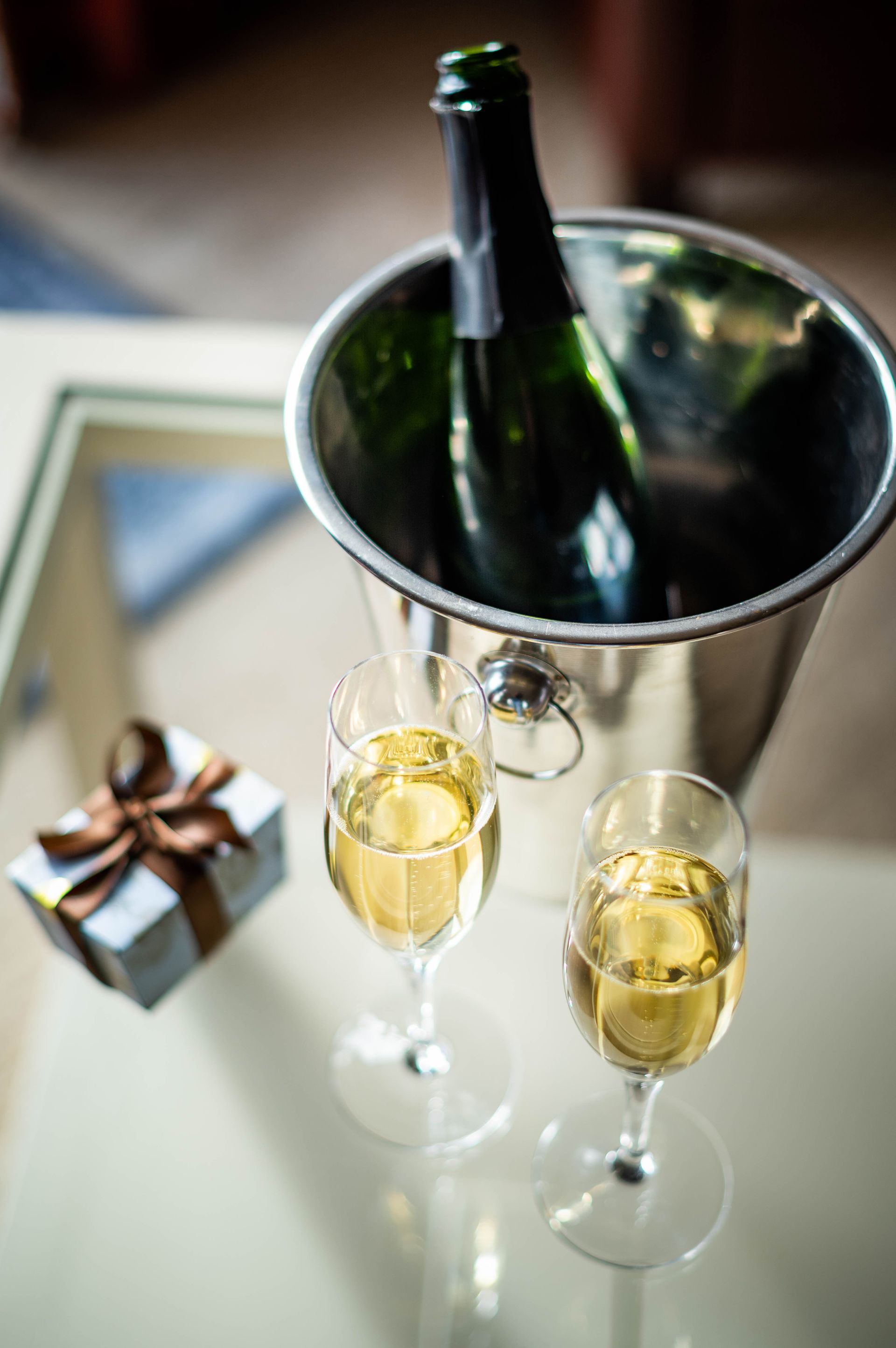 Two champagne flutes filled with bubbly, and a bottle of sparkling wine in an ice bucket on a glass table.