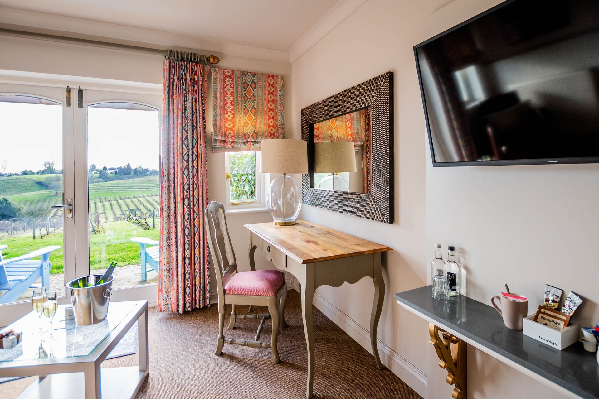 A hotel room overlooking a vineyard, featuring a desk, chair, mirror, and TV. Bright colors and natural light.