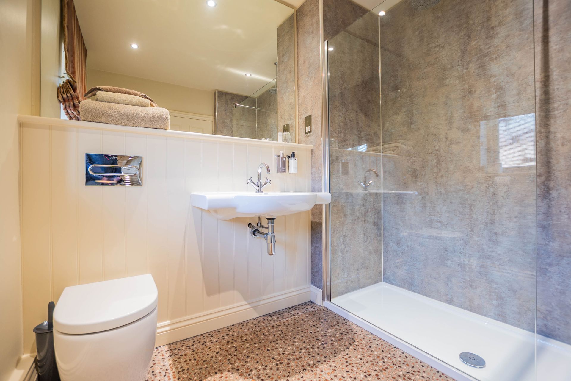 Bathroom with white toilet and sink, a glass-walled shower, and a speckled floor. Towels sit above the sink.