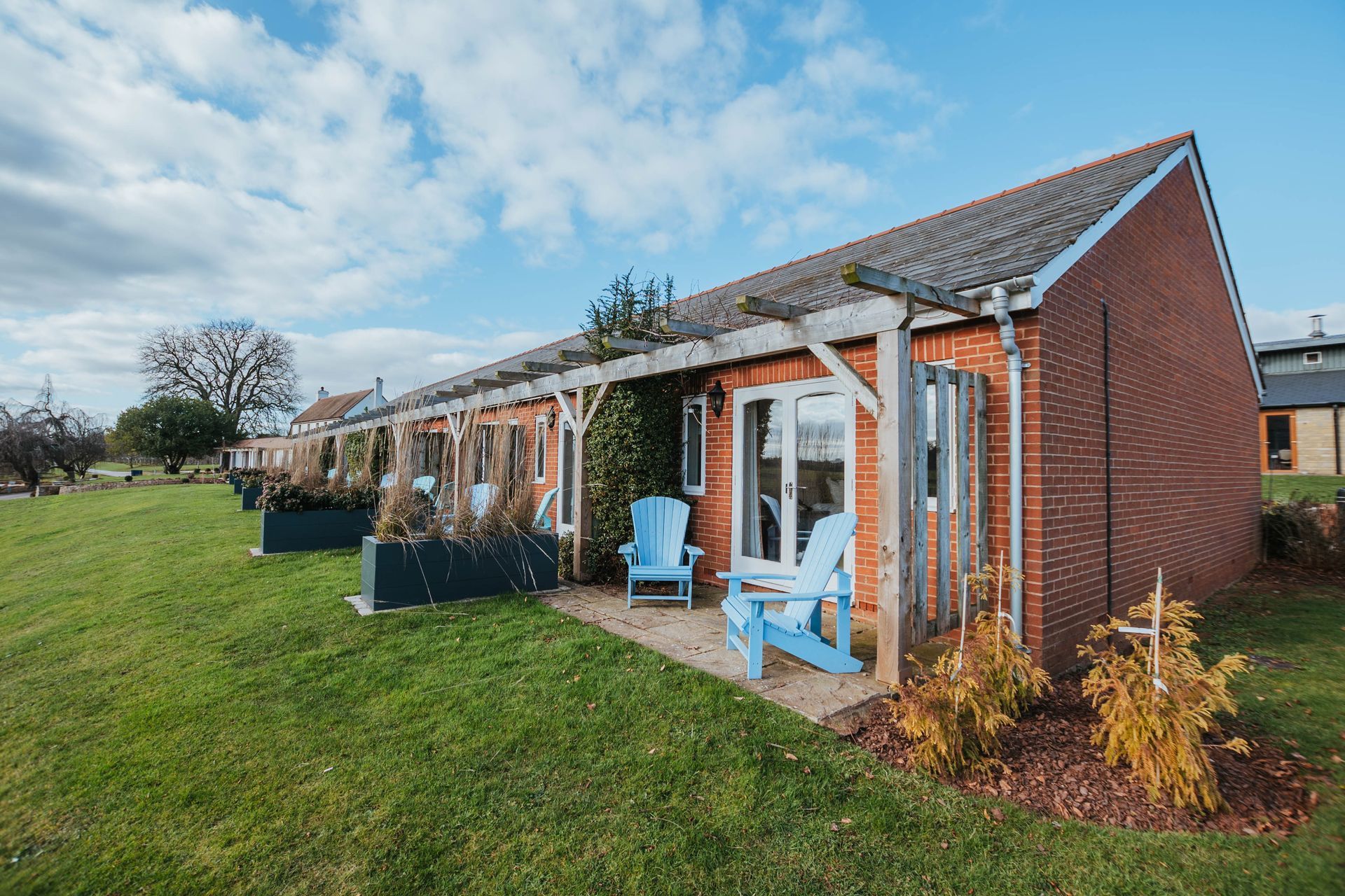 Brick cottages with seating on a grassy lawn under a cloudy sky. Two blue Adirondack chairs sit outside one cottage.