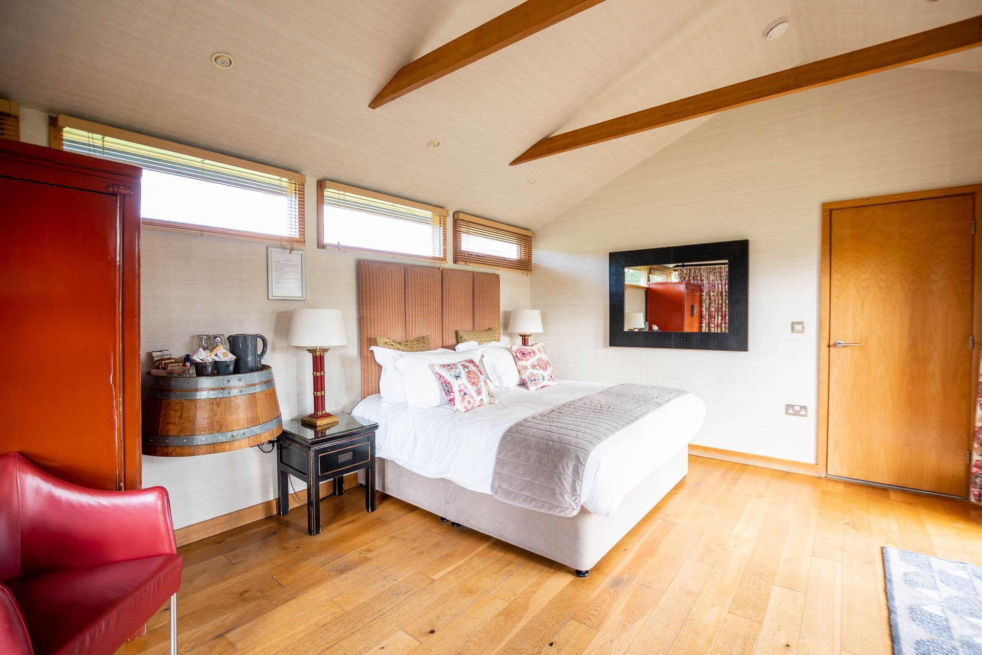 A bright bedroom with a bed, TV, and small windows. Features include a red armchair, wooden floors, and a decorative barrel.