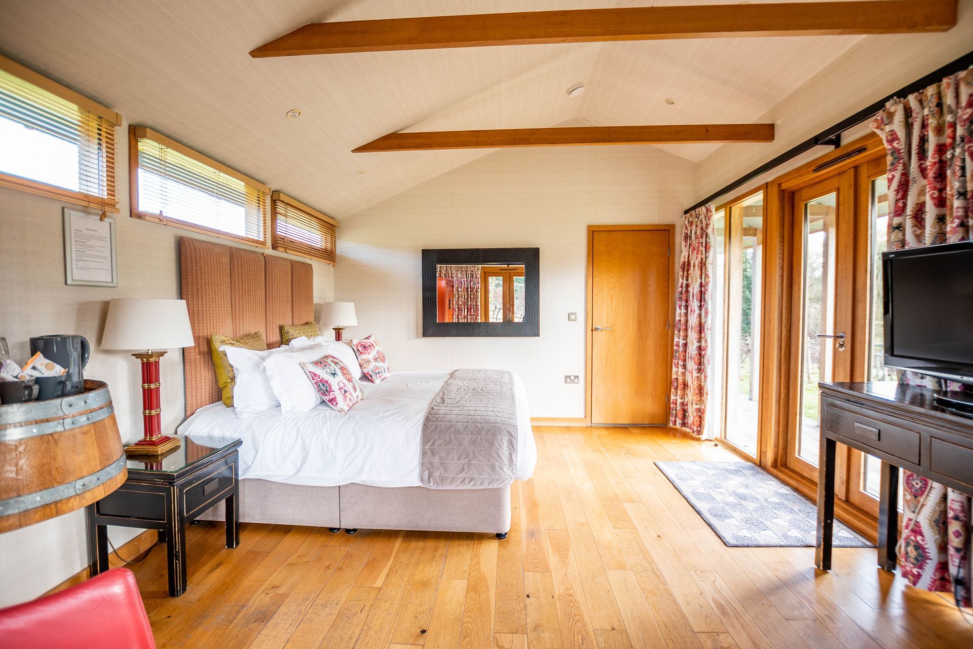 Bedroom with a bed, wooden floors, and French doors leading outside. The room has neutral walls, and brown wood beams with a tv on the wall.
