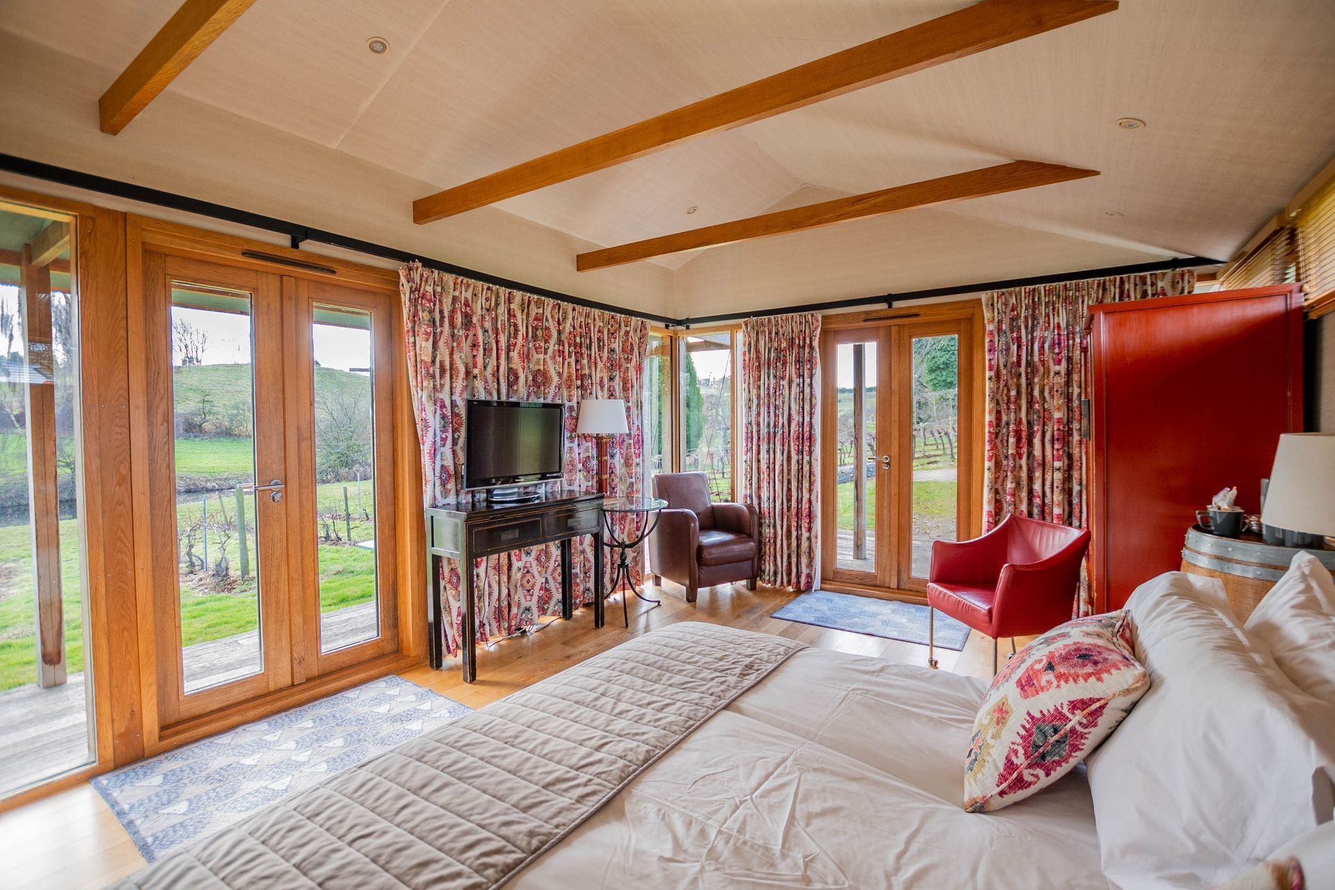 Bedroom with French doors overlooking greenery. Features a bed, TV, chairs, and patterned curtains.