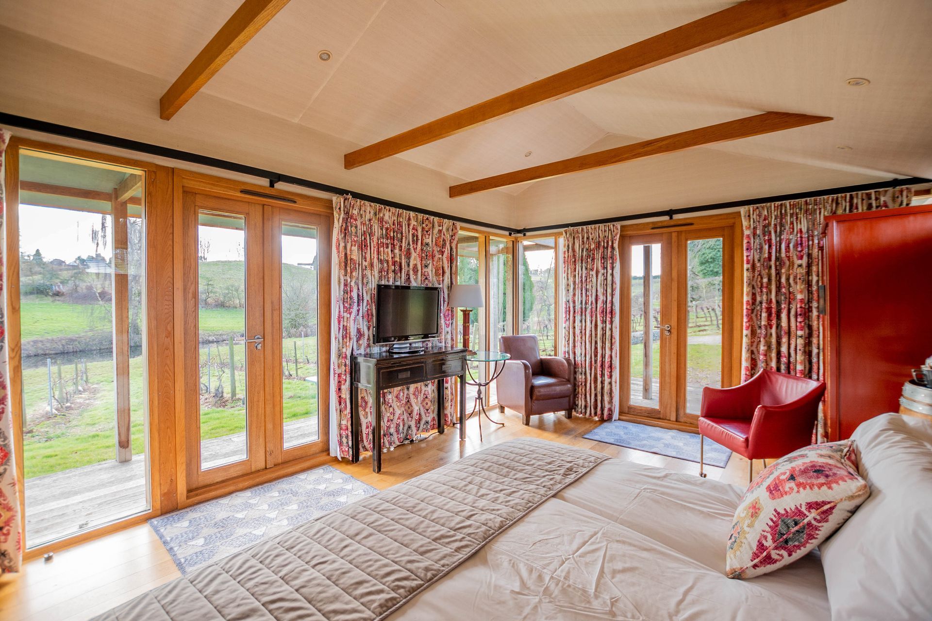 Bedroom with large bed, windows, and doors overlooking a green field. The room has wood beams, red accents, and patterned curtains.