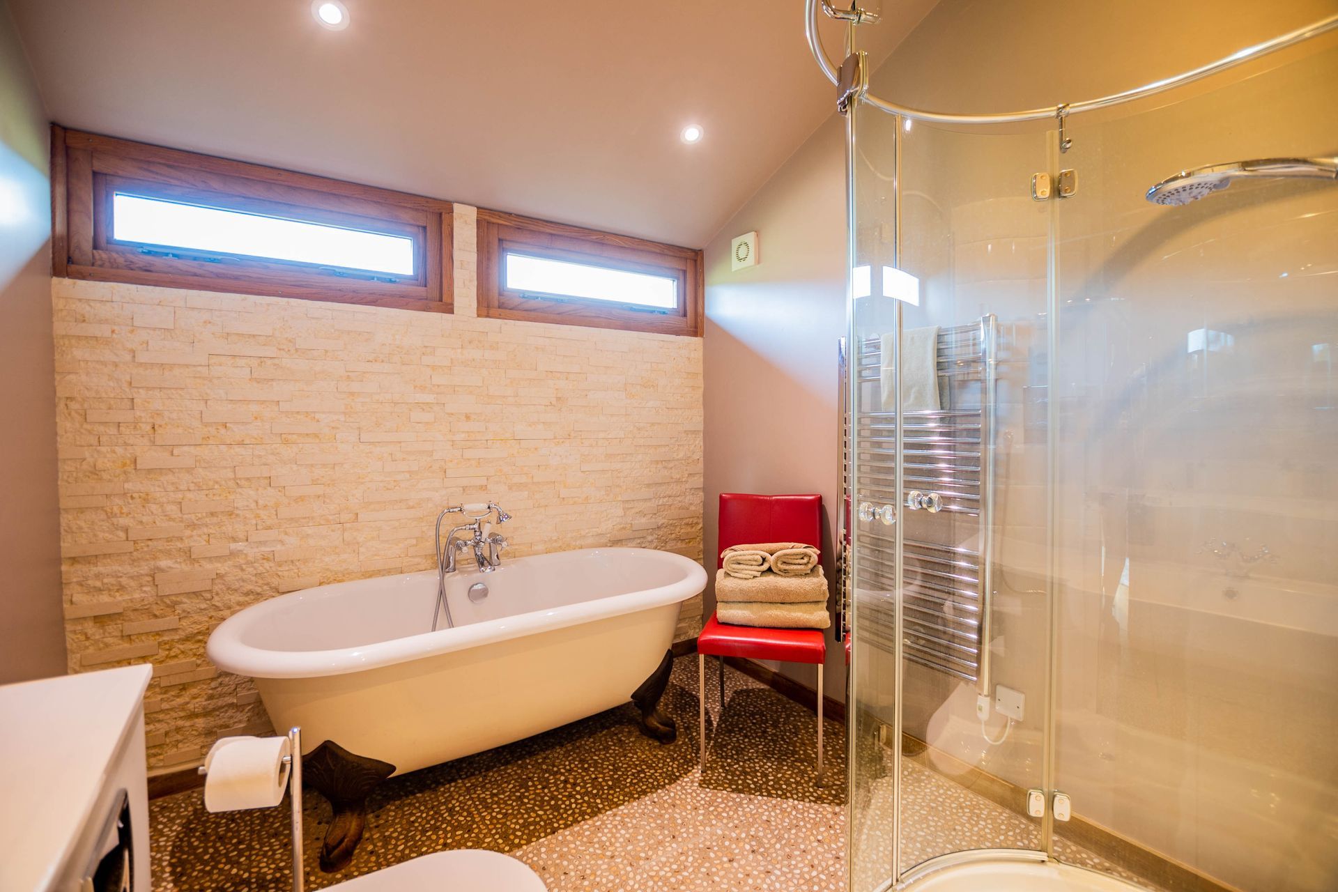 Bathroom with a white clawfoot bathtub, glass shower, red chair, and pebble flooring. Two windows are visible at the top.
