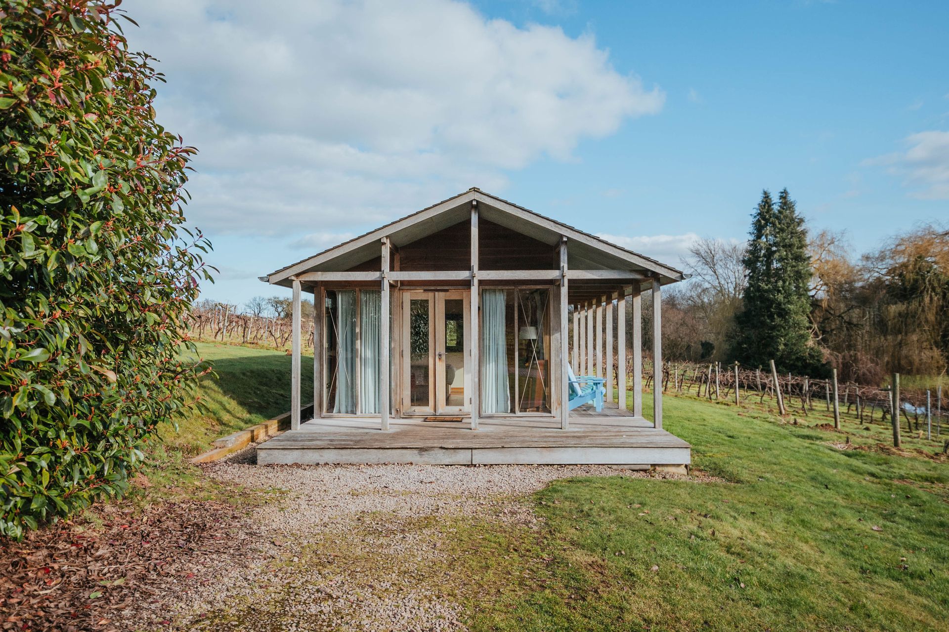 A small wooden cabin with glass doors and a porch sits in a grassy field with a vineyard and trees in the background on a sunny day.