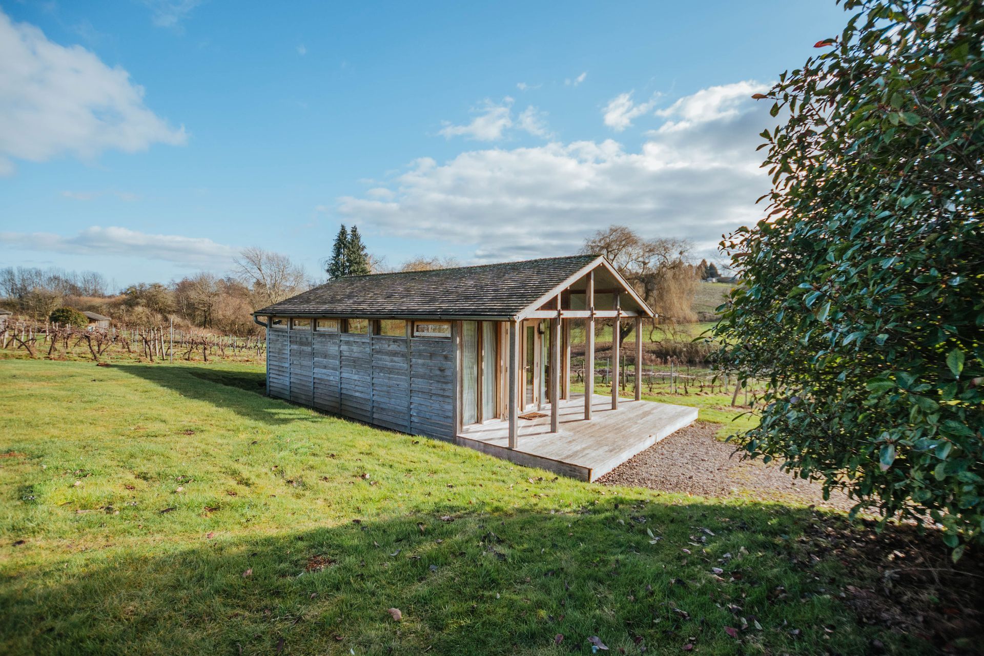 A small wooden structure with a porch in a grassy field, under a blue sky.  Vineyard visible in the background.