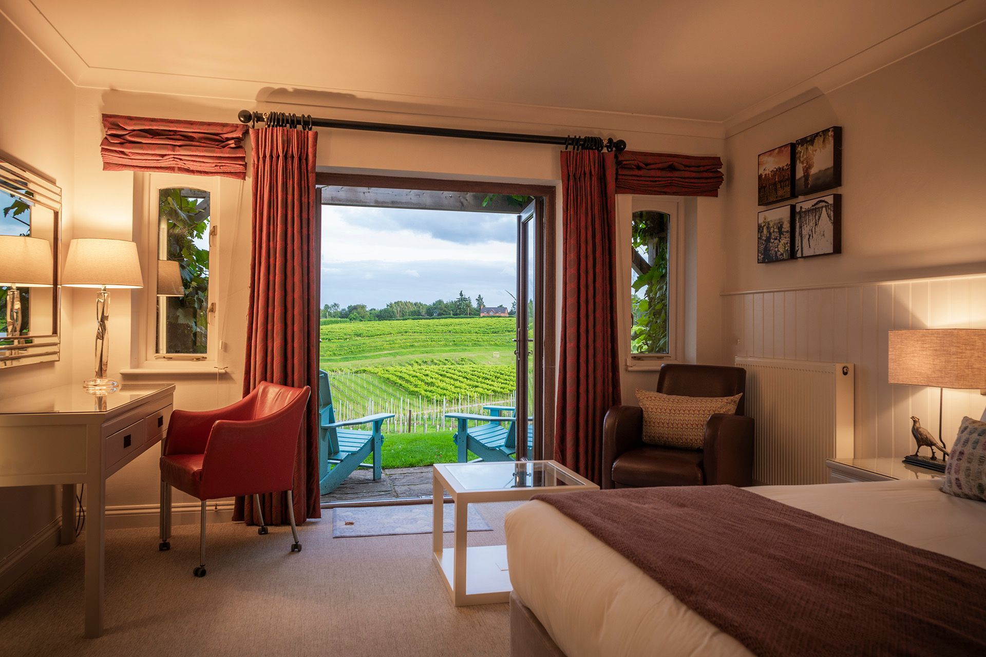 A hotel room with a view of a vineyard. The room has a bed, desk, and opens onto a patio with chairs.