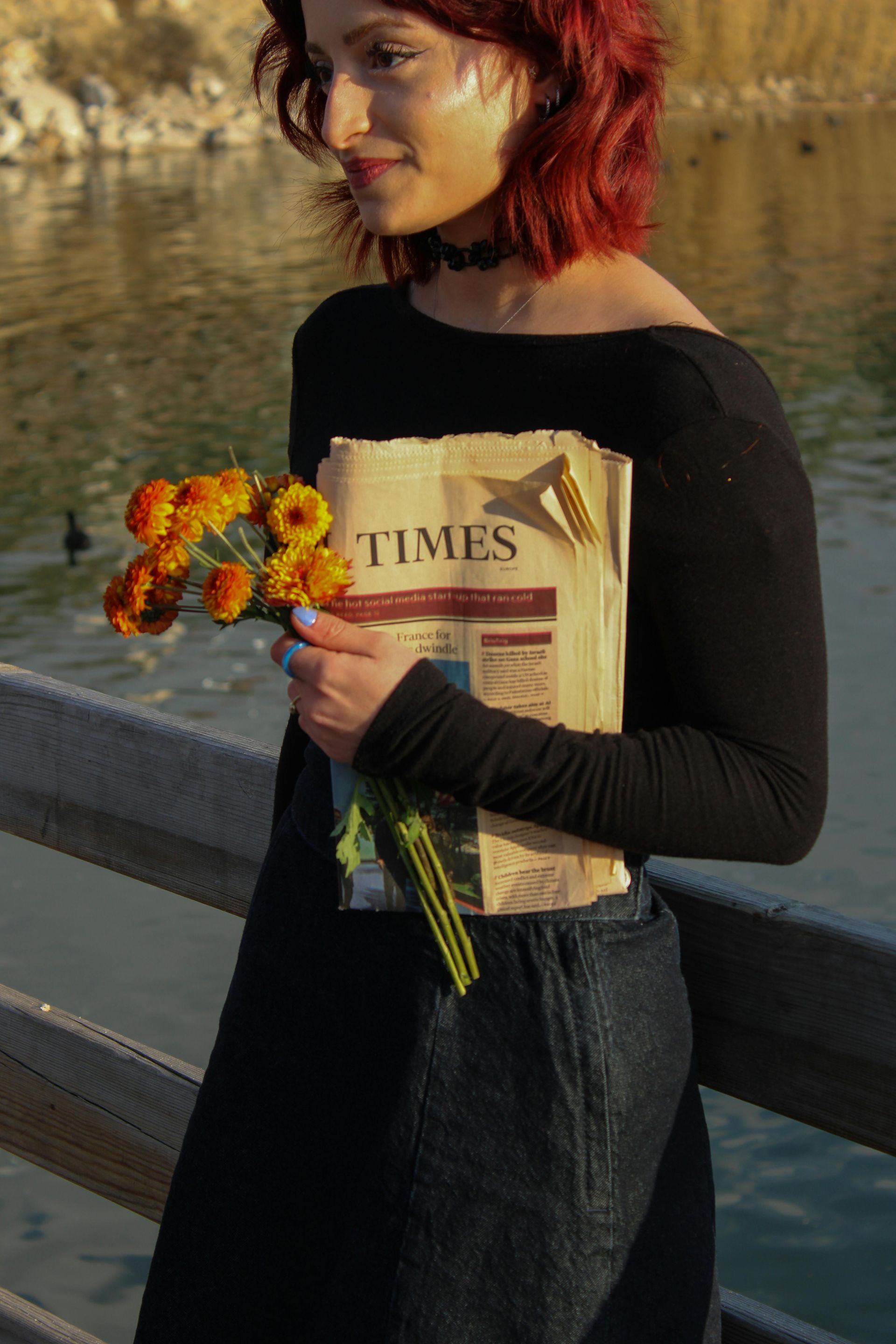 Woman with red hair holds flowers and a newspaper on a wooden bridge over water, wearing a black top and jeans.
