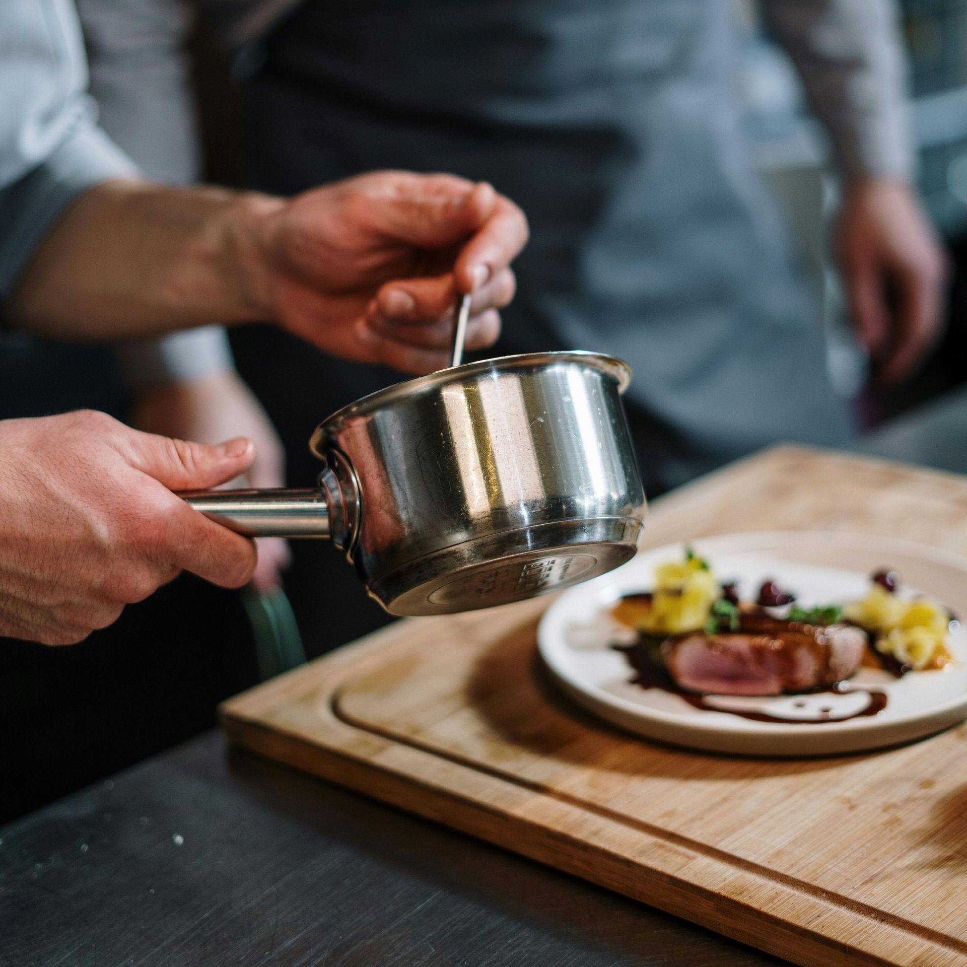 Chef plating a dish, drizzling sauce onto food on a white plate atop a wooden cutting board. A person wearing a gray apron stands in the background.