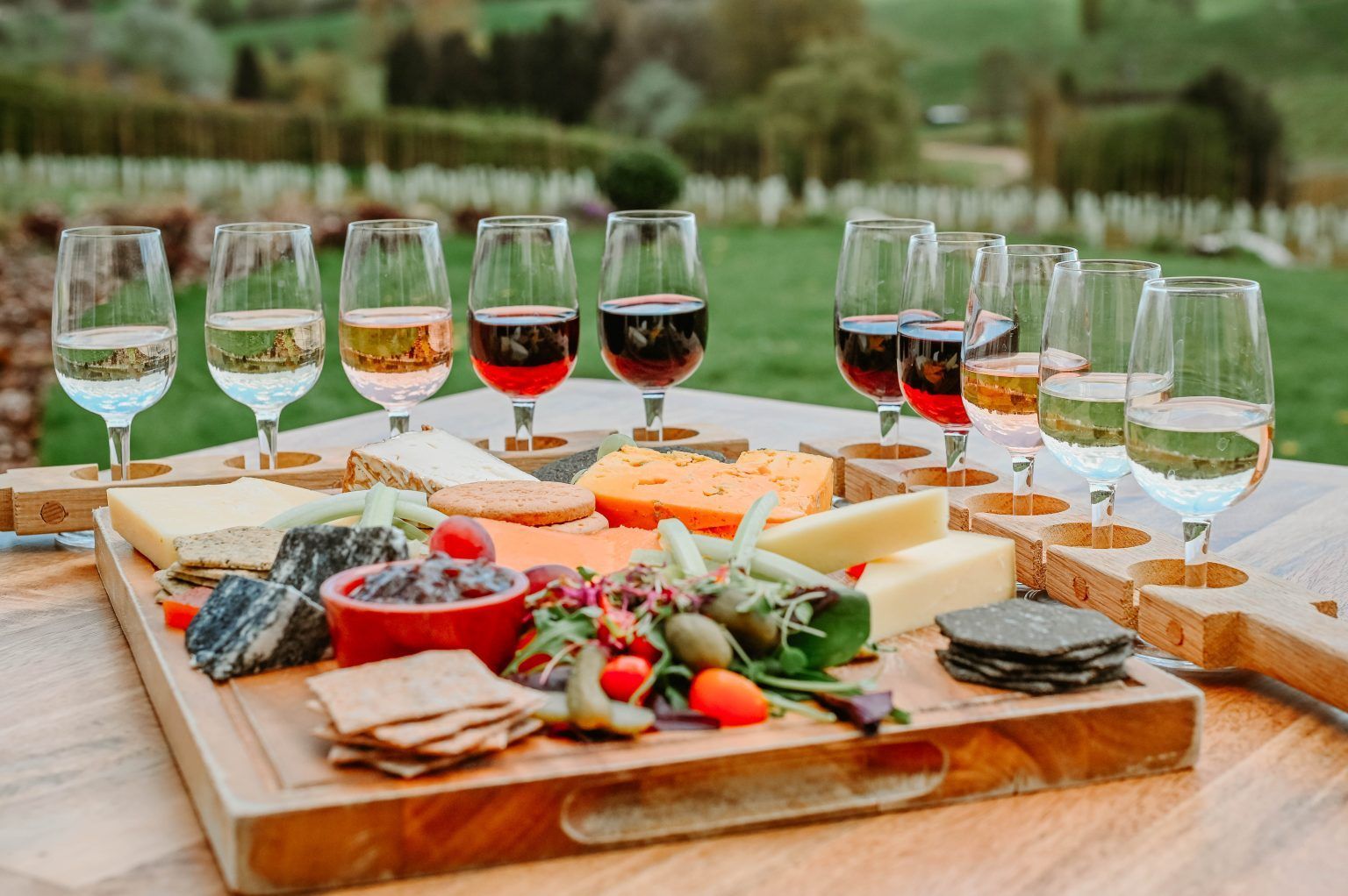 A wooden board with various cheeses, crackers, and olives is paired with wine glasses. Outdoors in a vineyard setting.