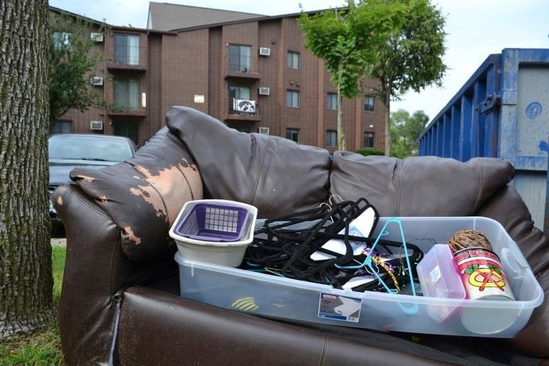 A brown leather couch is filled with junk in front of a dumpster.
