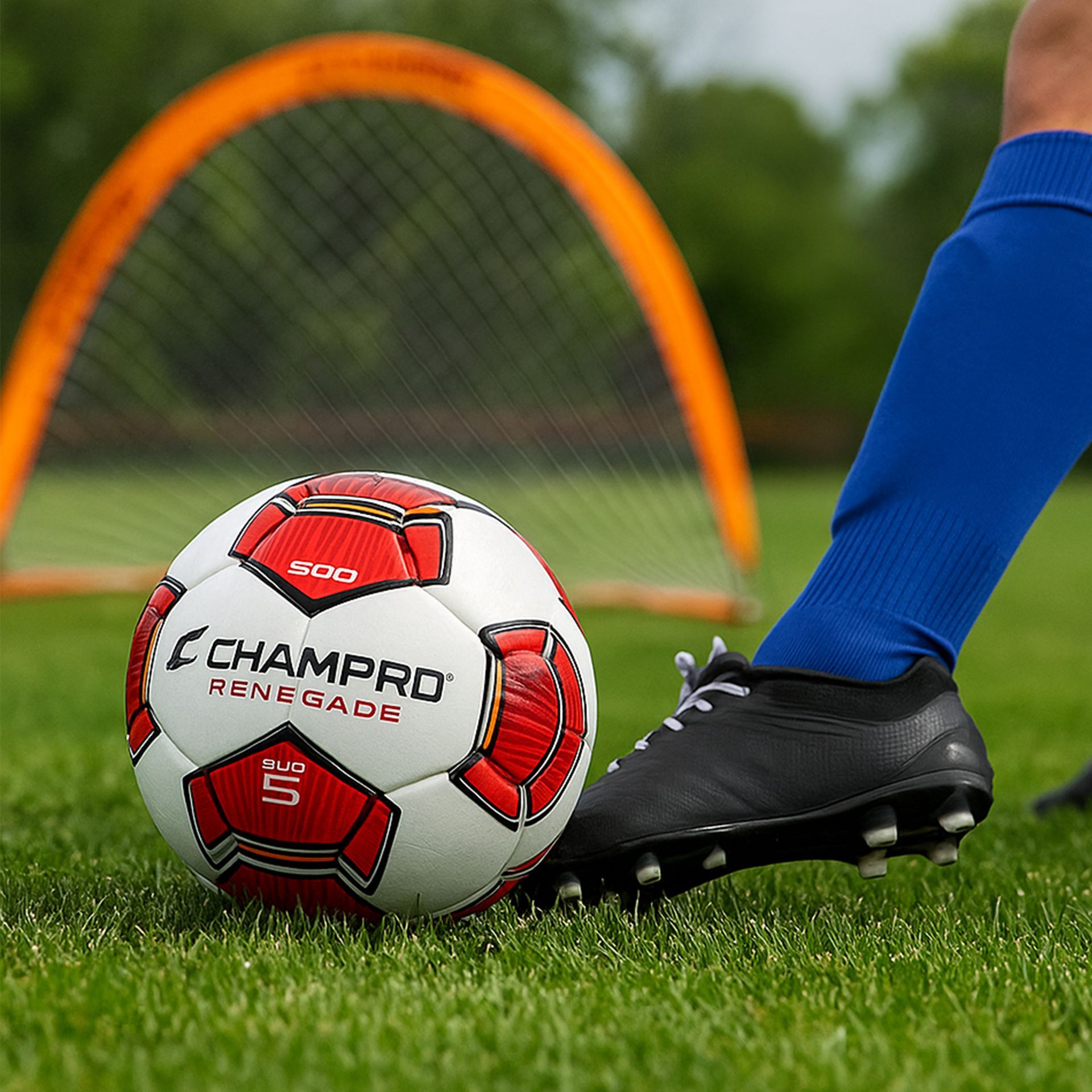 Soccer ball on grass, foot kicking. Orange goal in background, blue sock.