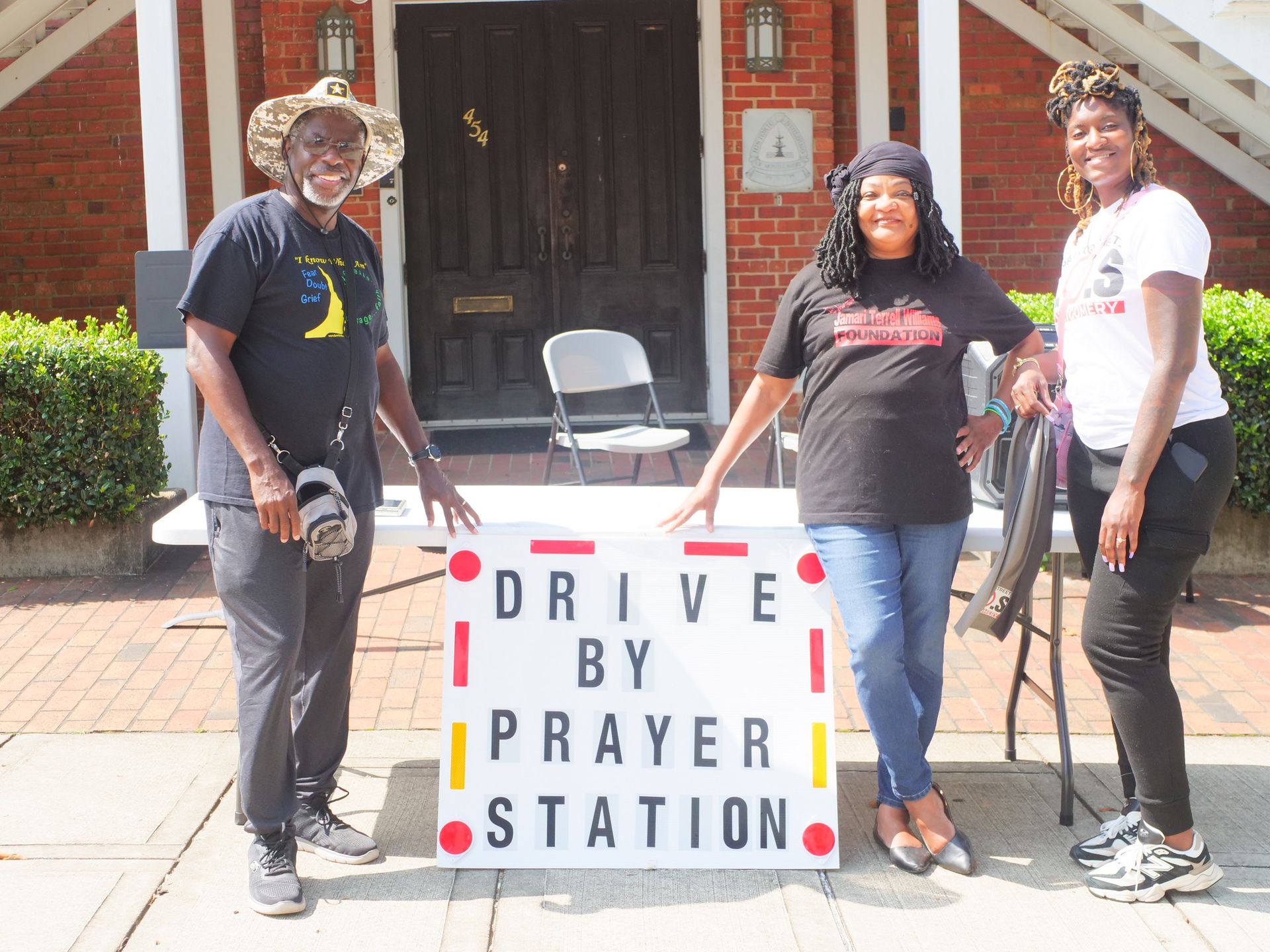 Three people are standing in front of a sign that says drive by prayer station