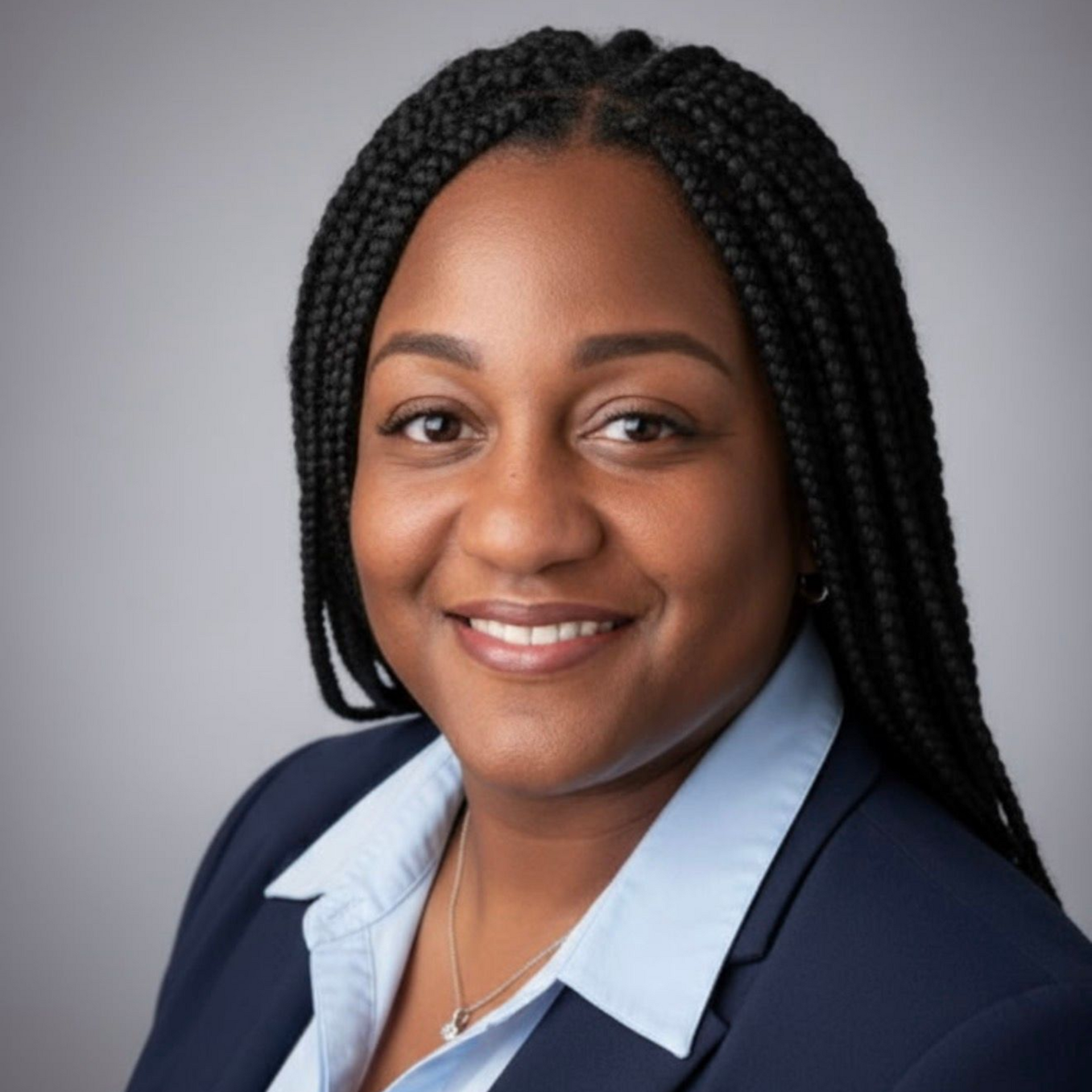 Woman smiling, wearing a blue blazer and light blue shirt. Braided hair, studio backdrop.