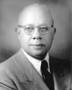 A black and white photo of a bald man wearing glasses and a suit and tie.