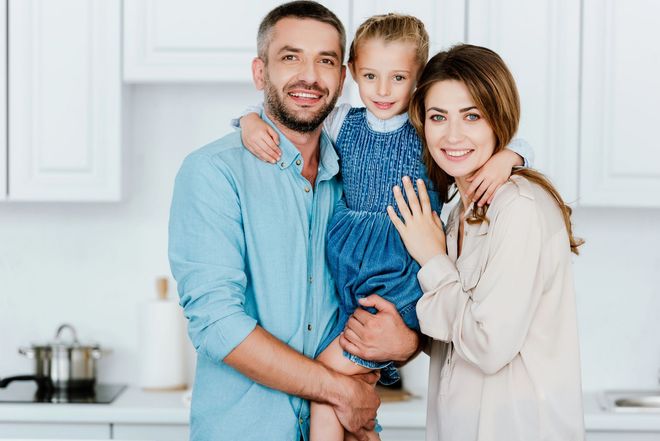 Una famiglia sorridente di tre persone posa in una cucina luminosa, con un uomo che tiene in braccio un bambino accanto a una donna.