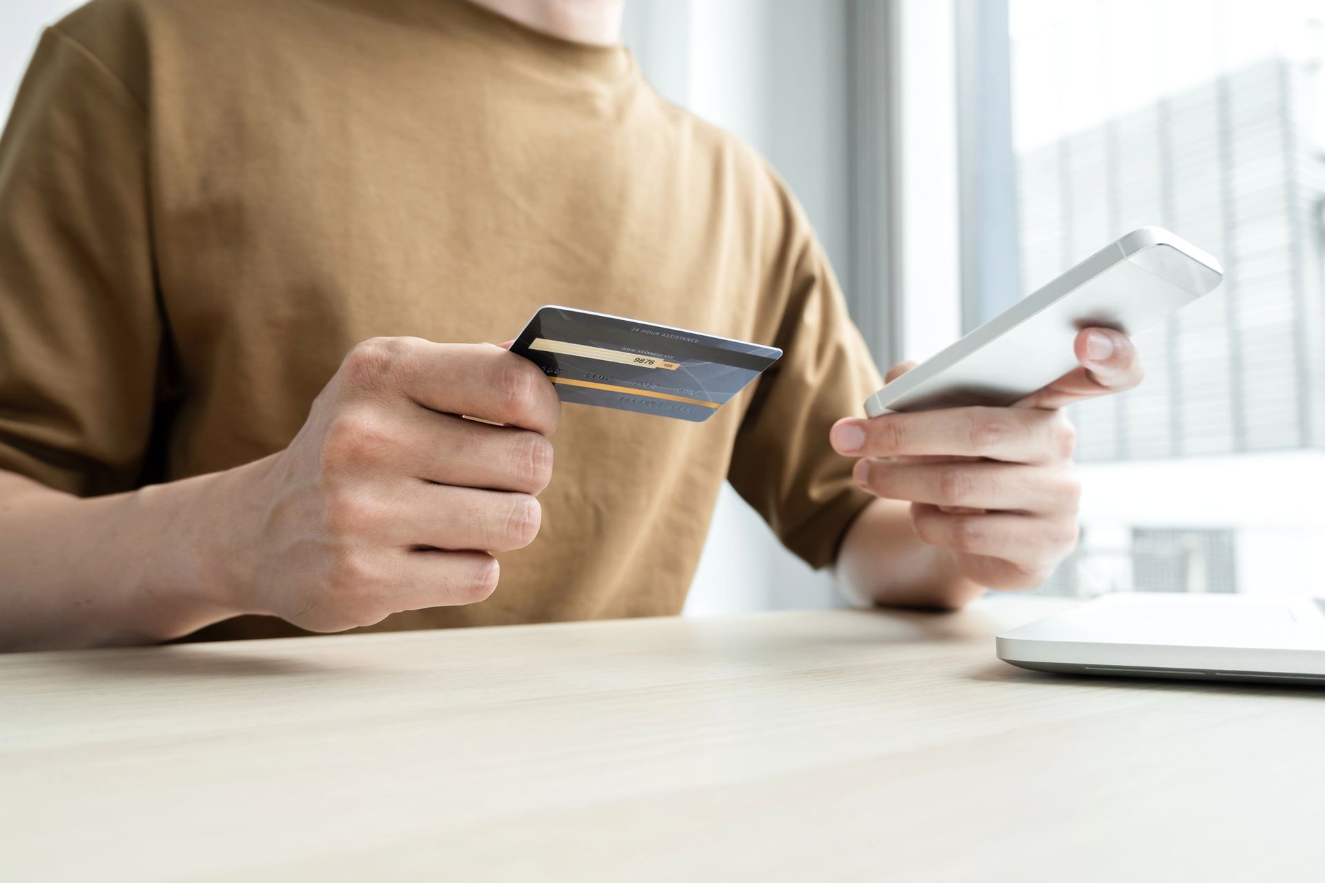 Una persona, seduta a una scrivania, tiene in mano una carta di credito e uno smartphone, mentre effettua un pagamento online.