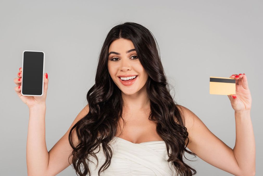 Donna sorridente che tiene in mano uno smartphone e una carta di credito su uno sfondo grigio uniforme.