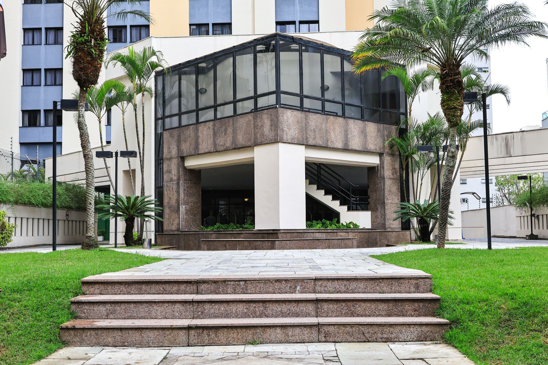 Entrada do apartamento com degraus, passarela coberta e palmeiras em frente ao prédio.