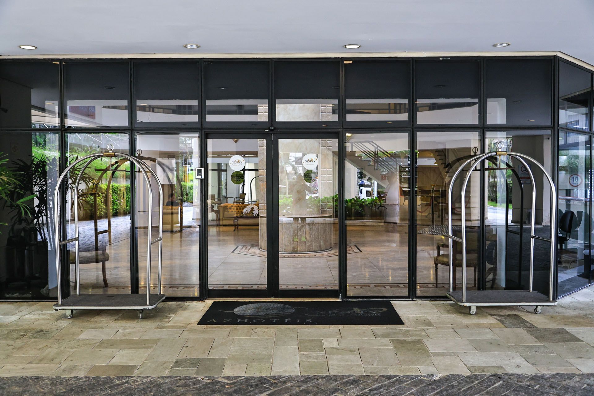 Entrada do edifício com fachada de vidro, carrinhos de bagagem de metal e um capacho preto sob um toldo.