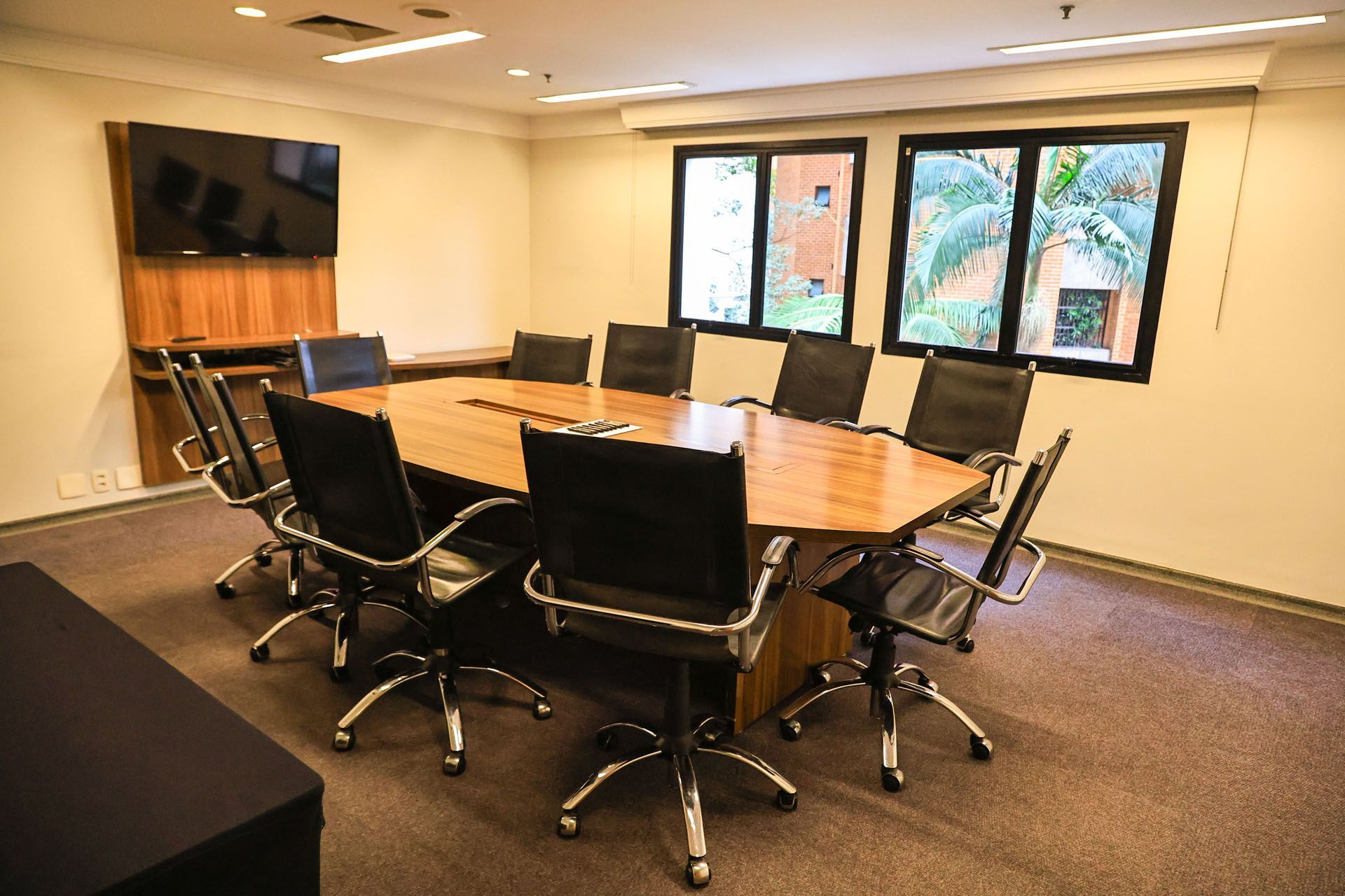 Sala de conferências moderna com uma grande mesa oval, cadeiras pretas, TV de parede e janelas com vista para as árvores.