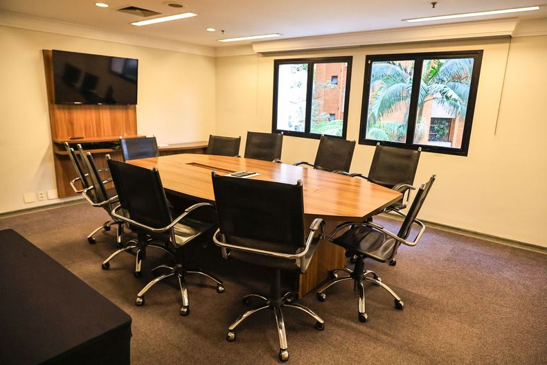 Sala de conferências moderna com uma grande mesa oval, cadeiras pretas, TV fixada na parede e janelas que permitem a entrada de luz natural.