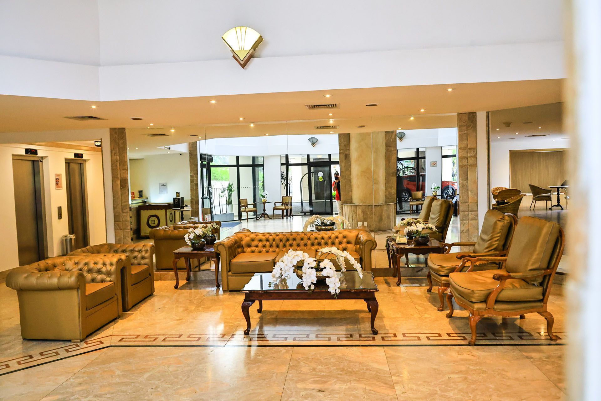 Hall de entrada do hotel com sofás bege, mesas de centro e uma área de entrada envidraçada e iluminada.